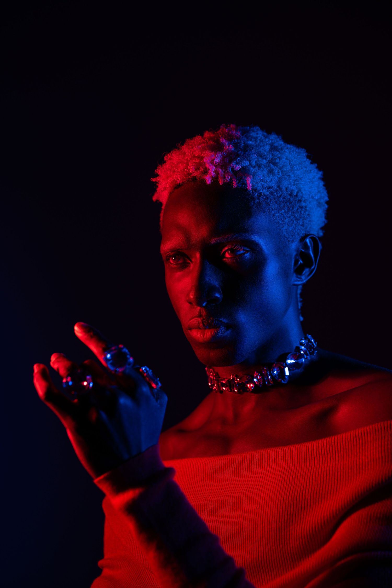 Dramatic portrait of person with blonde cropped hair illuminated by red and blue neon lights.