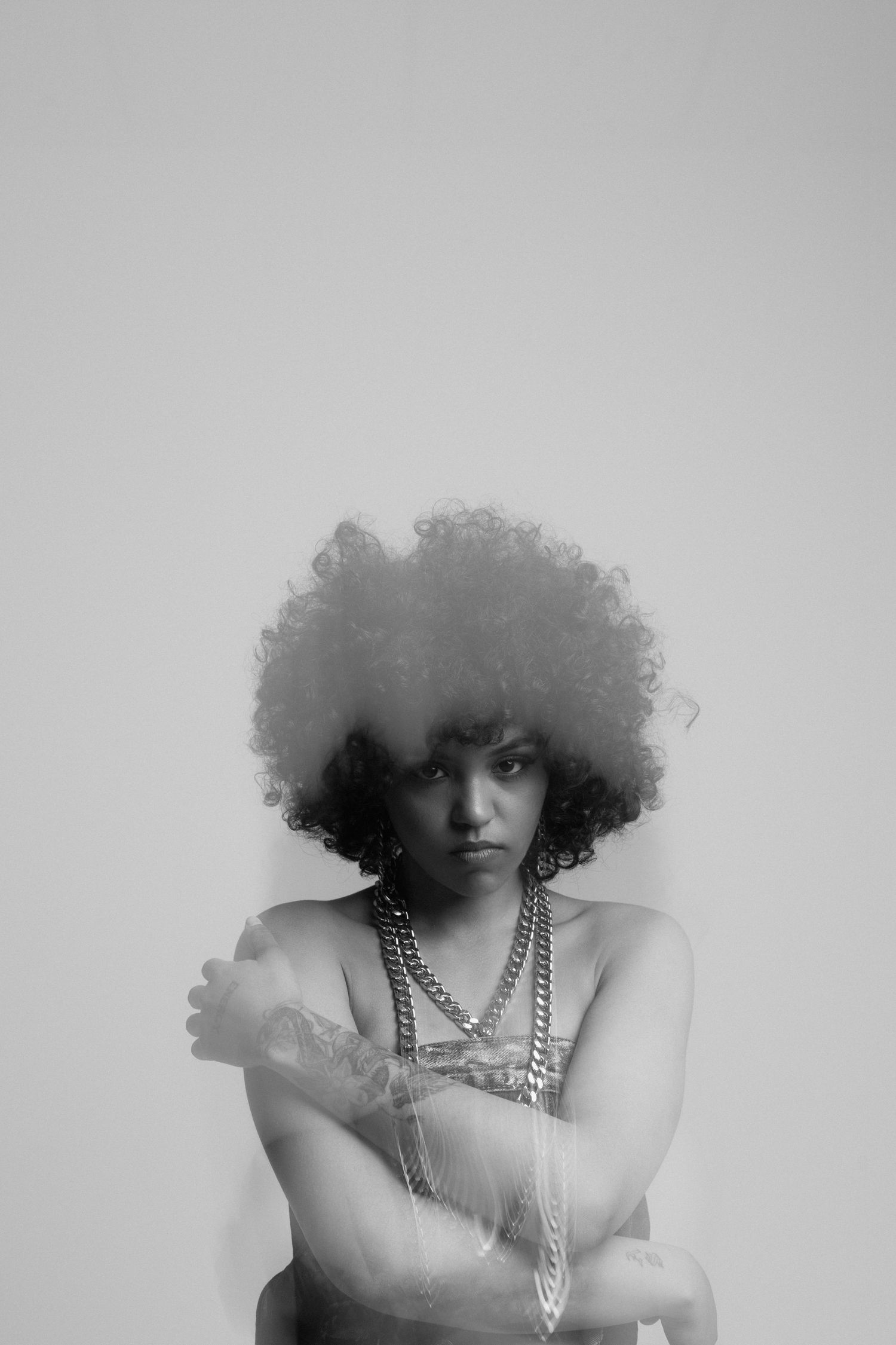 Black and white portrait of a woman with afro hair wearing chain necklaces, arms crossed, studio shot.