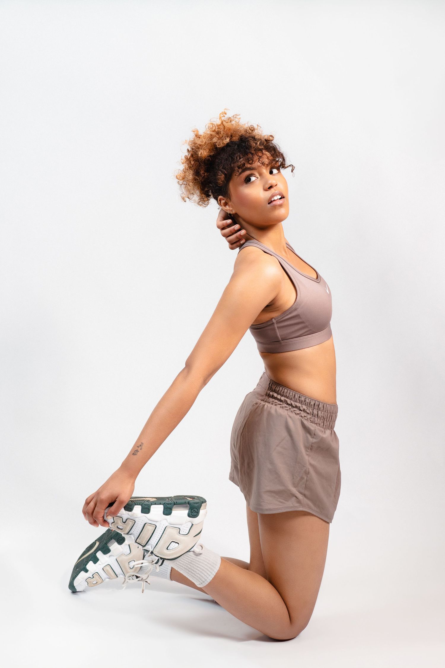 Athletic woman in taupe Nike sports bra and shorts kneeling while holding sneaker on white background.
