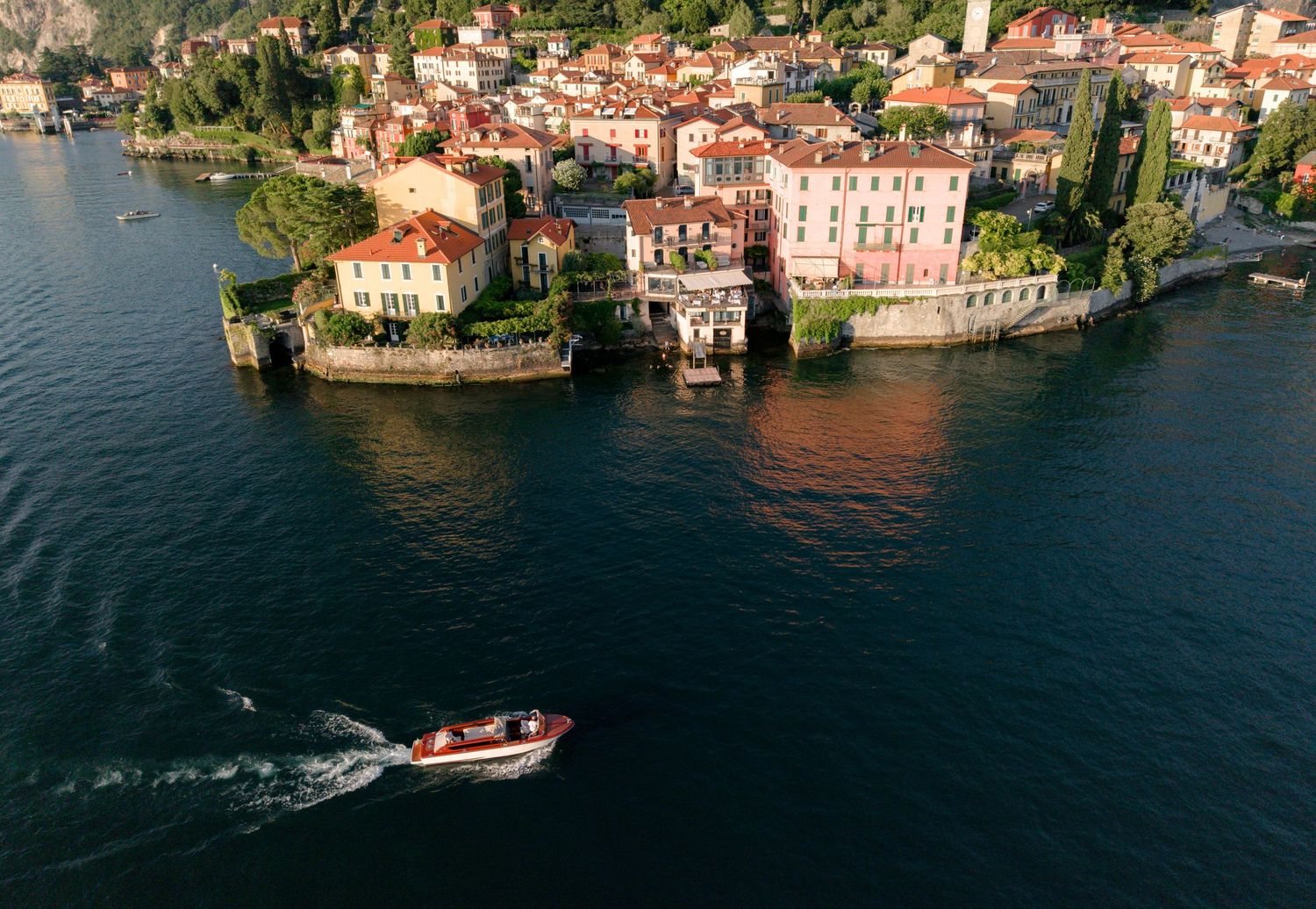 Aerial view of a picturesque Italian lakeside village with colorful buildings and a speedboat on deep blue water.