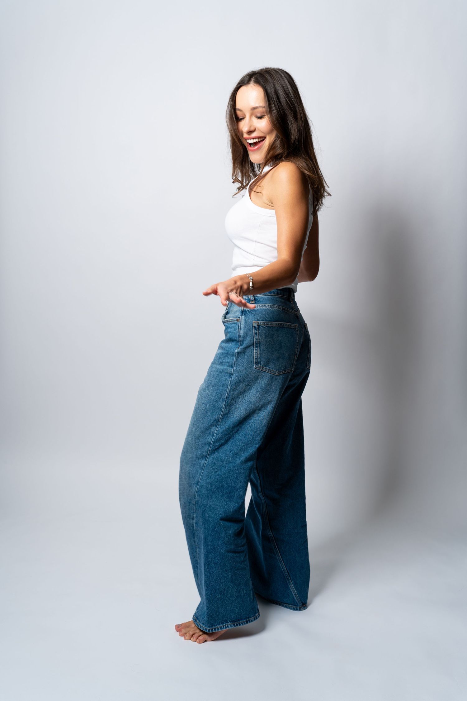 Side profile of smiling woman in white tank top and wide-leg blue jeans posing in studio.