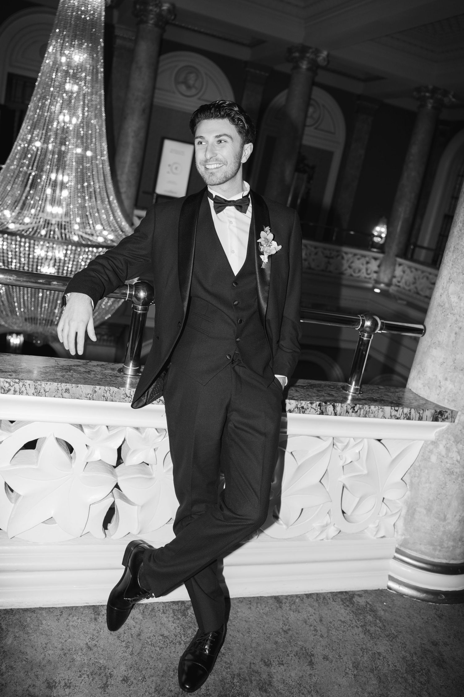 Elegant groom in black tuxedo with bow tie and boutonniere leaning against ornate marble railing in grand venue.