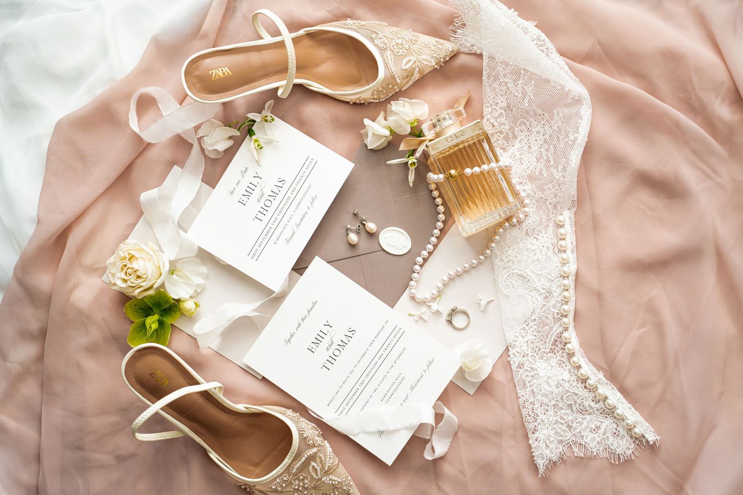 Elegant wedding flat lay with invitations, bridal shoes, pearl jewelry, and lace accessories on blush fabric.