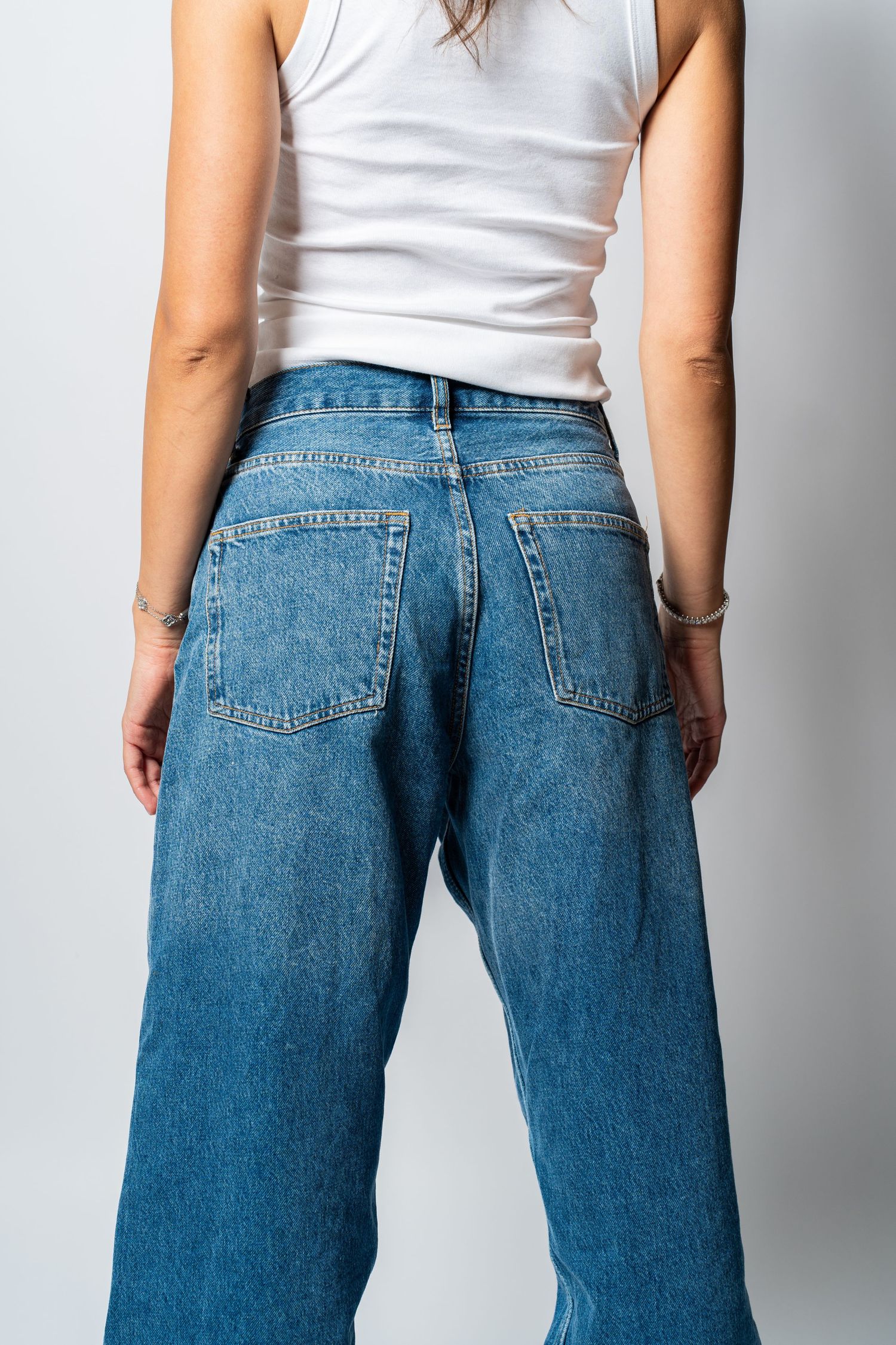 Back view of woman wearing wide-leg blue denim jeans and white tank top against gray background.