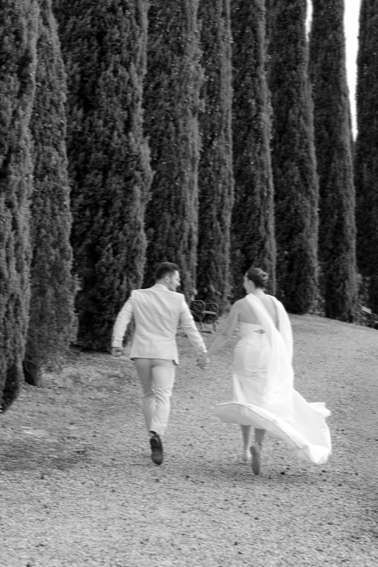 Black and white photo of bride and groom running hand in hand along cypress tree lined path.