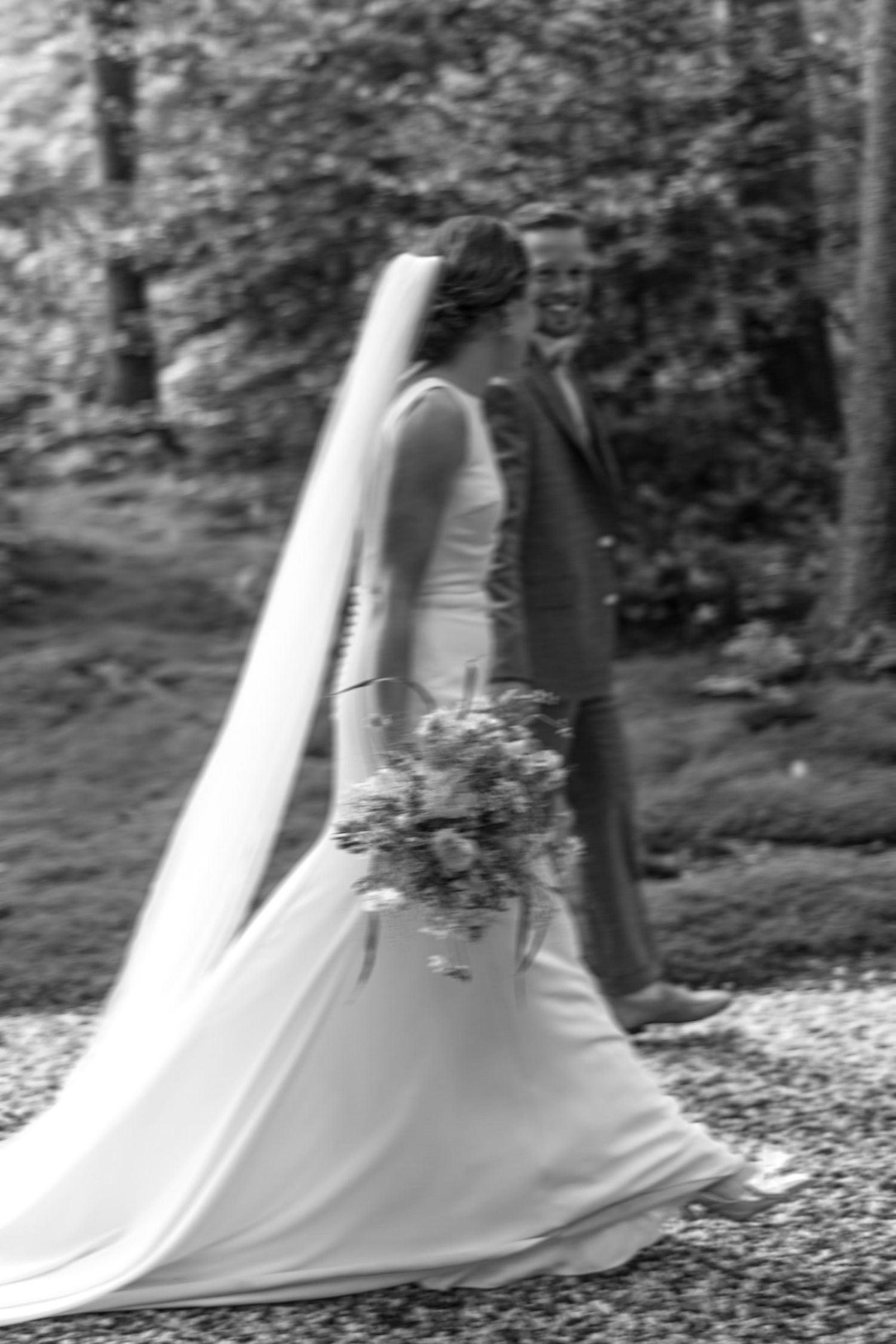 Black and white photo of a bride in a flowing gown and veil holding a bouquet with a smiling groom outdoors.