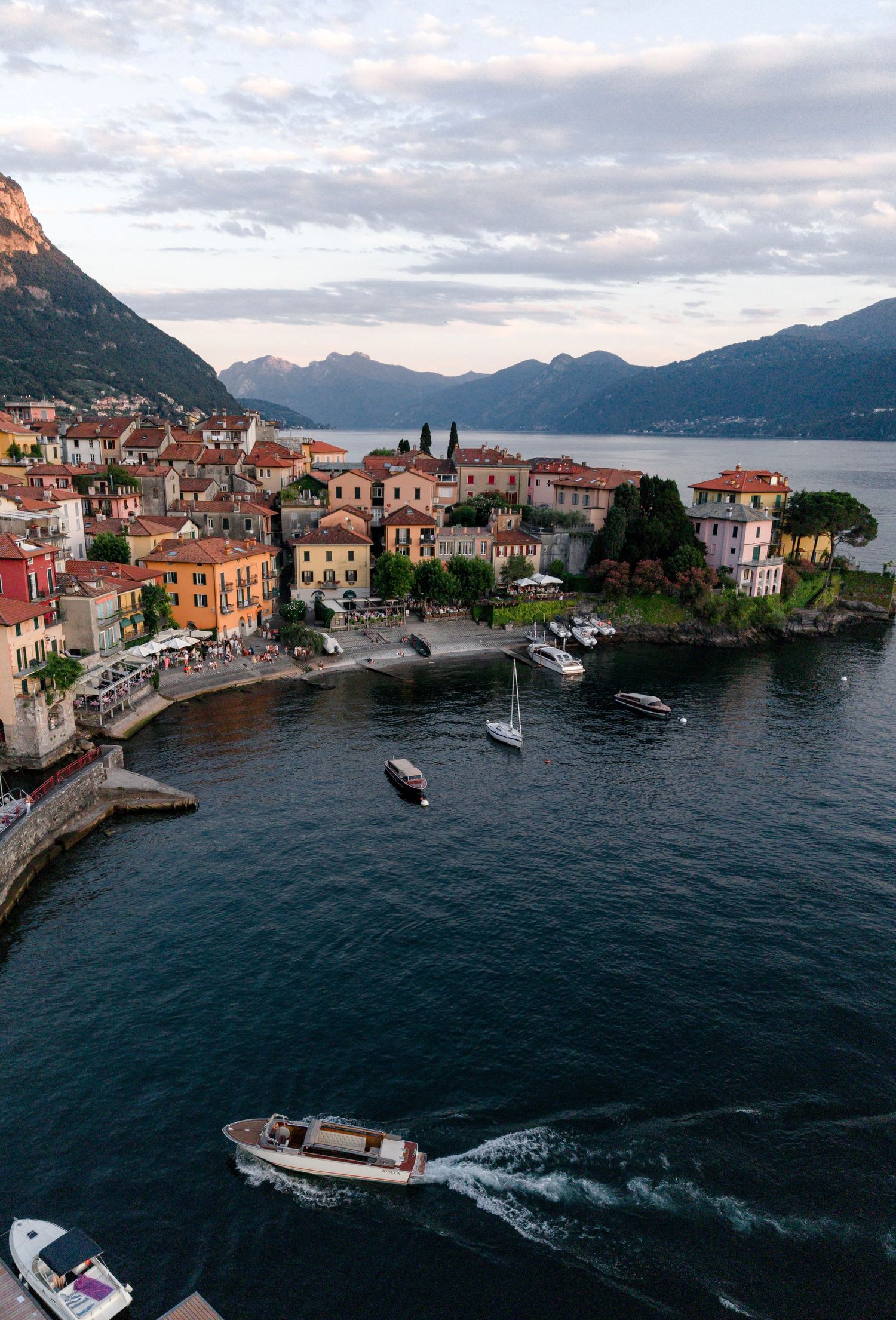 Aerial view of a picturesque Italian lakeside village with colorful buildings, boats on calm water, and mountains at dusk.