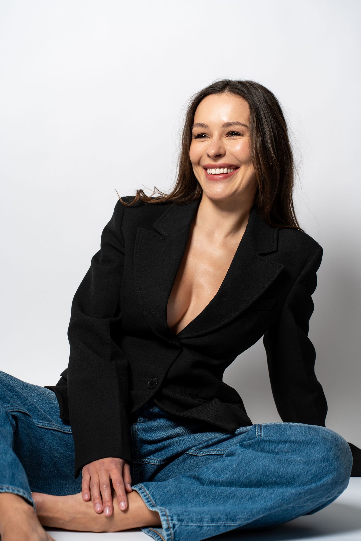 Laughing woman in black blazer and jeans sitting cross-legged on floor in studio portrait.