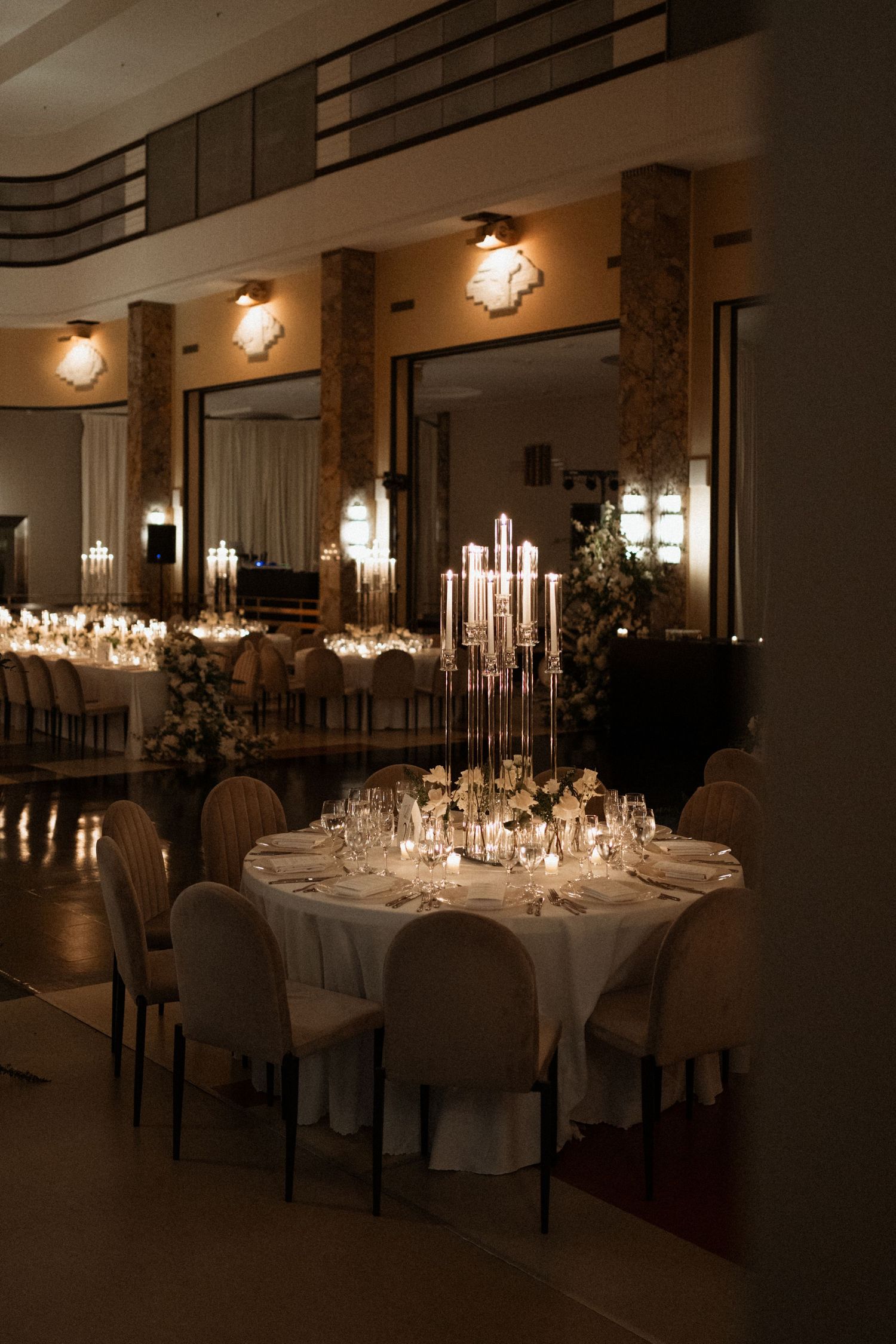 Elegant wedding reception tables set with tall candelabras and white linens in a dimly lit ballroom with wall sconces.