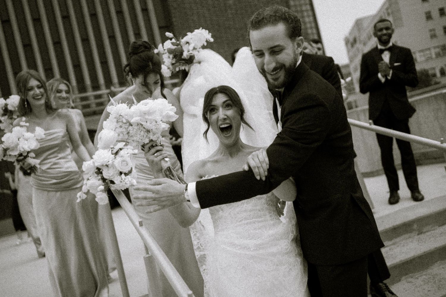 Black and white photo of a joyful bride and groom exiting a church, surrounded by bridesmaids holding bouquets.