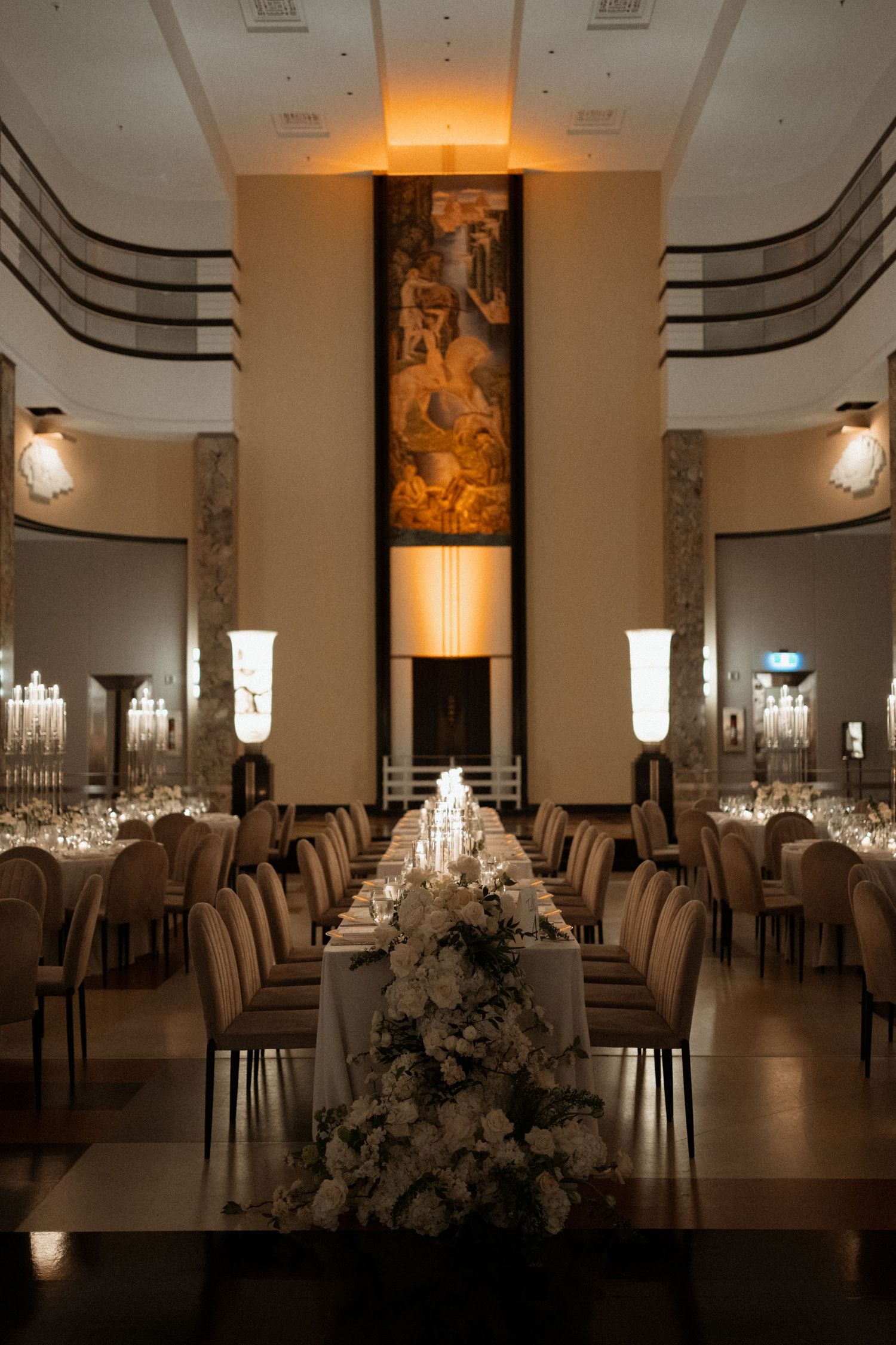 Elegant art deco ballroom with long banquet table adorned with white floral arrangements and ambient lighting.