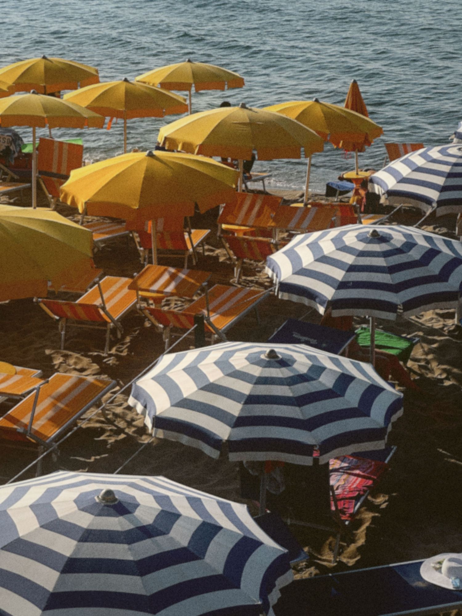 Yellow and striped beach umbrellas line a rocky Mediterranean coastline with blue waters in the background.