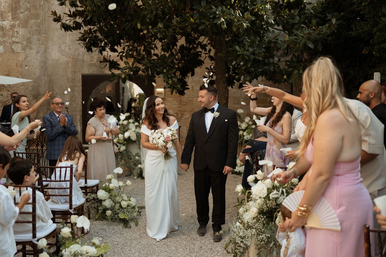 Wedding party walks through an outdoor garden venue with elegant floral arrangements and white chairs set up for ceremony.