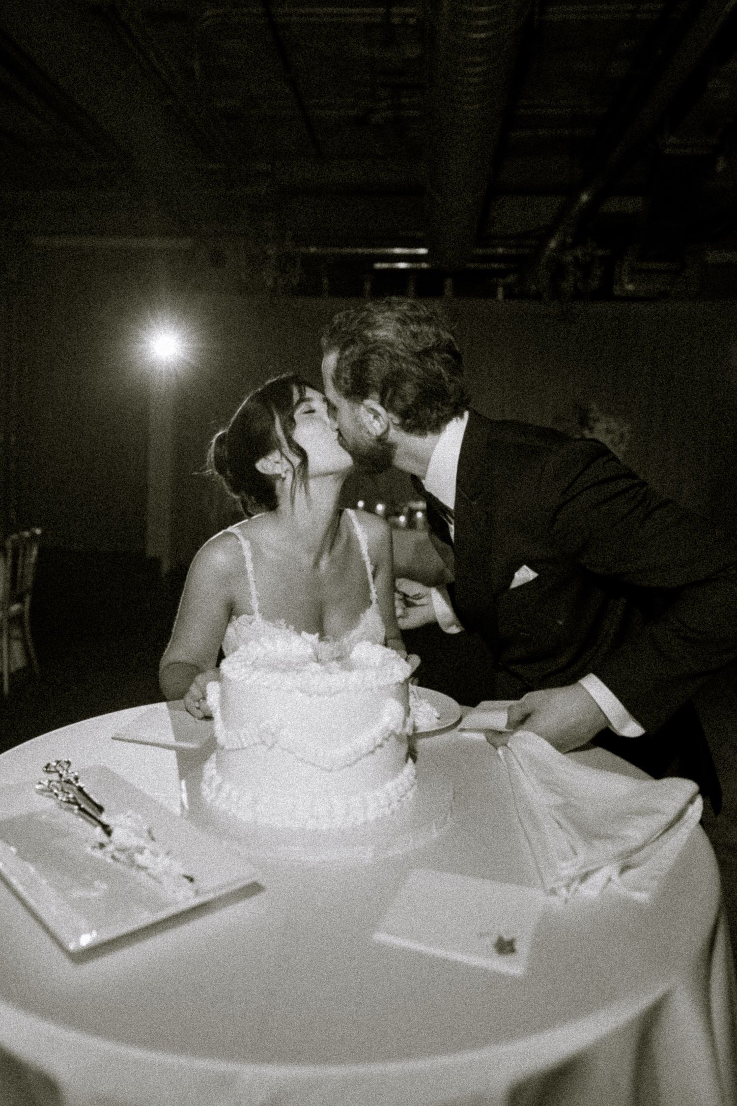 Black and white photo of bride and groom sharing a kiss while cutting their two-tier white wedding cake.