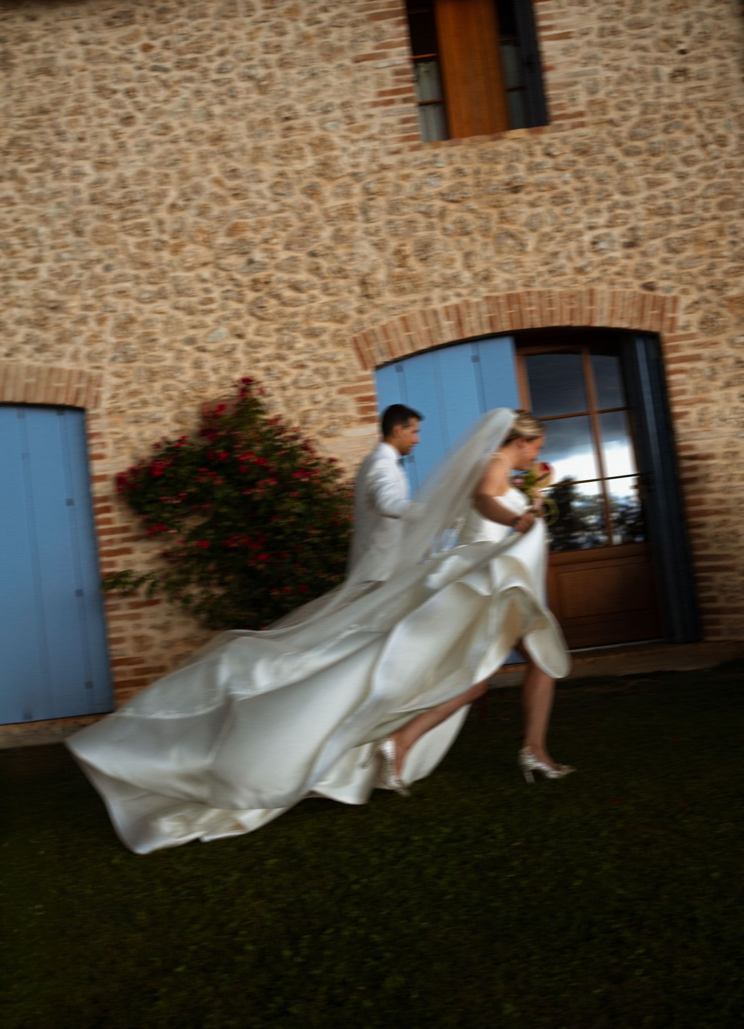 Bride in flowing white gown and groom walk along green lawn beside rustic stone building with blue shutters.
