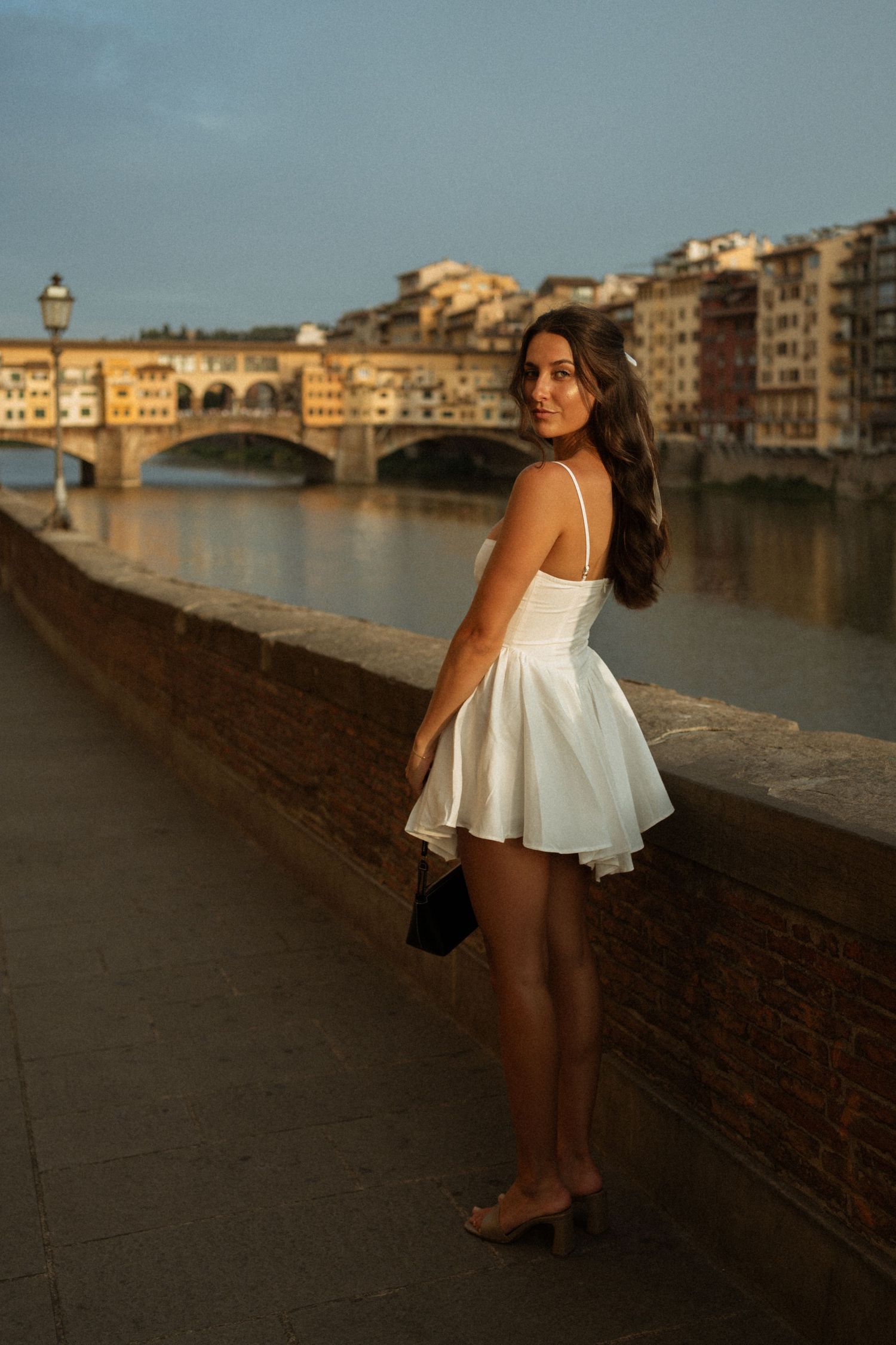 Woman in white dress poses by Arno River with Ponte Vecchio bridge in Florence, Italy at golden hour.