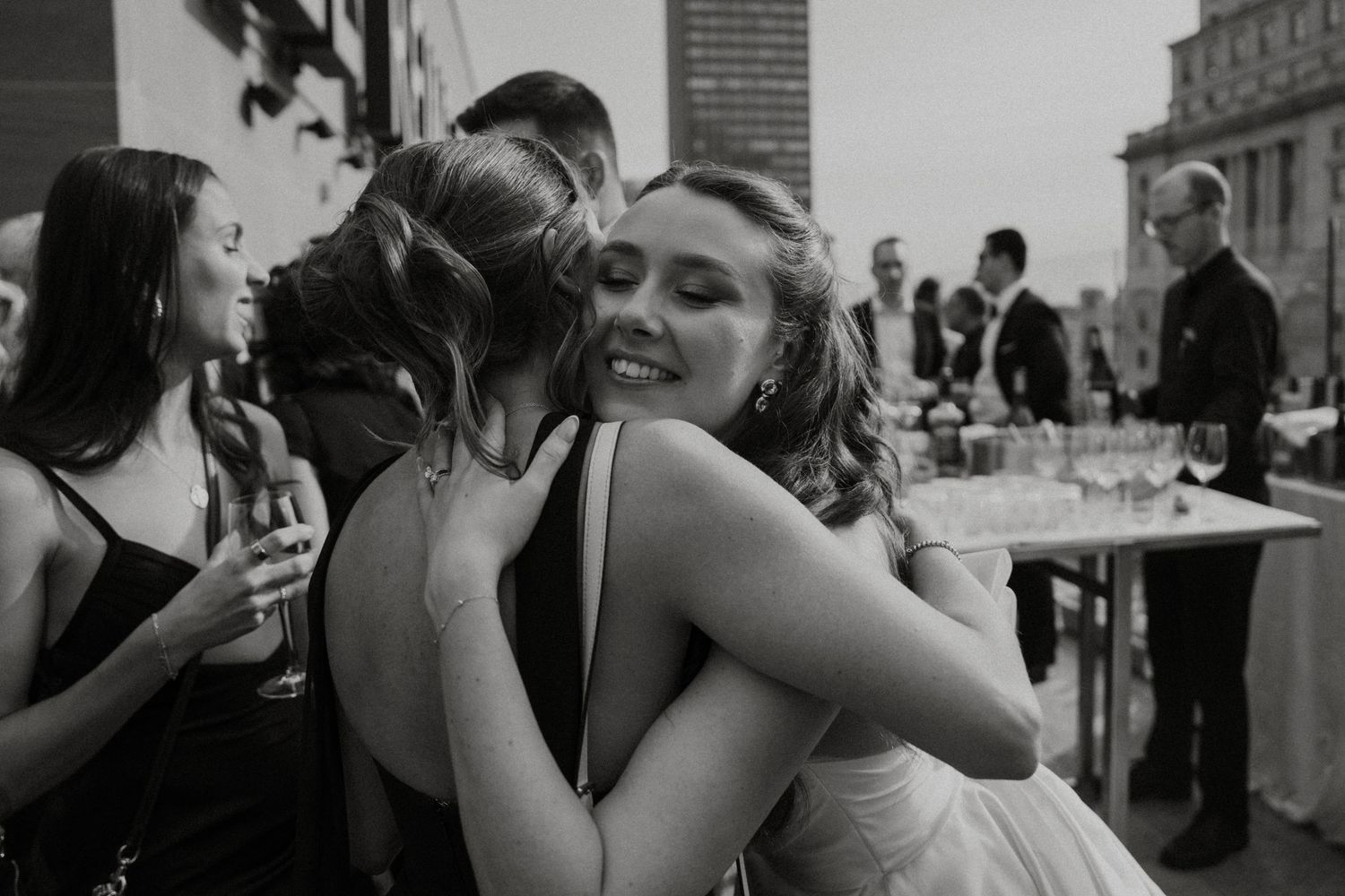 Two people share a warm embrace at an elegant black and white wedding reception with guests in the background.