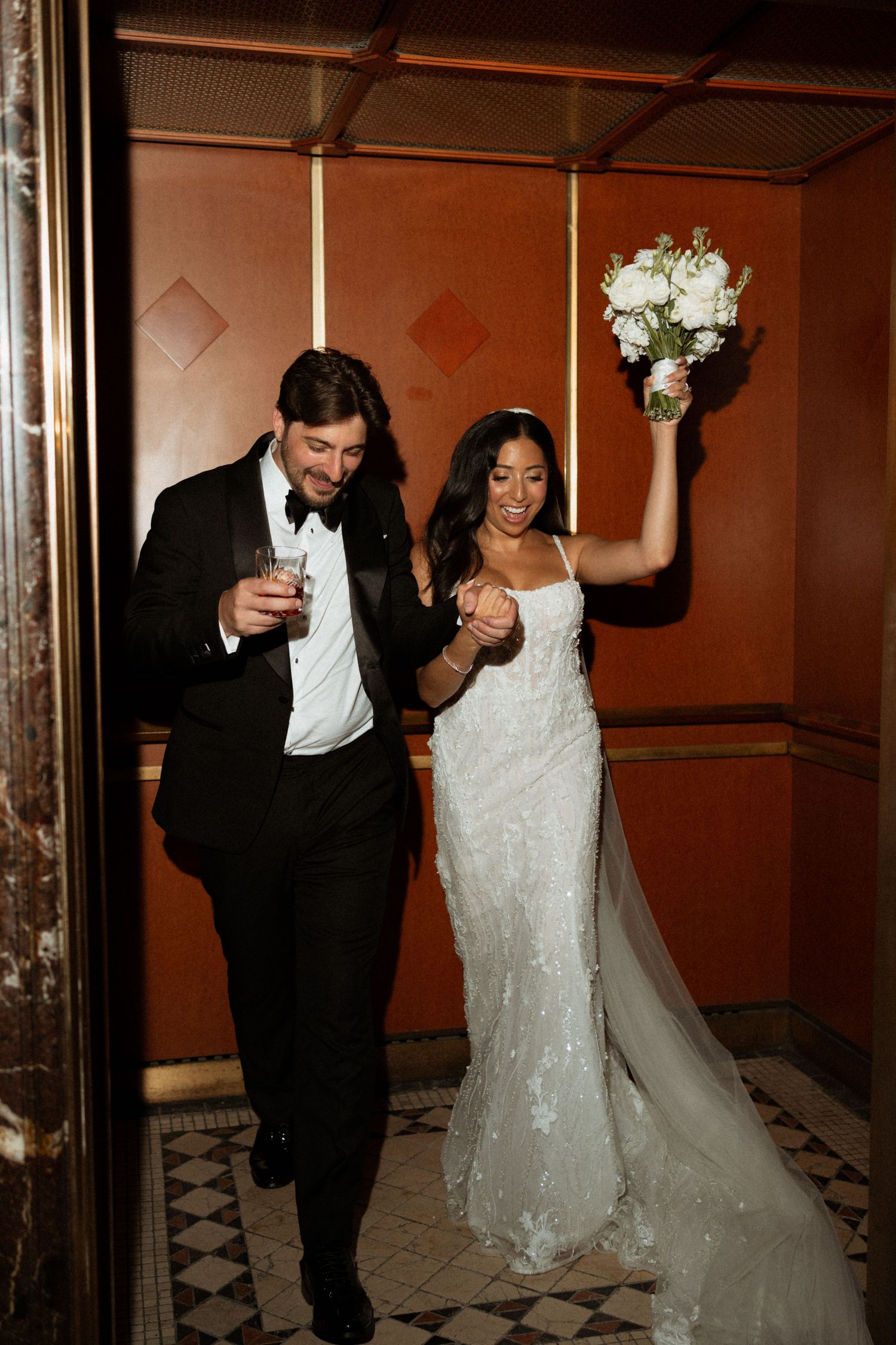 A bride and groom celebrate joyfully while walking through an elegant hallway with checkered floors.