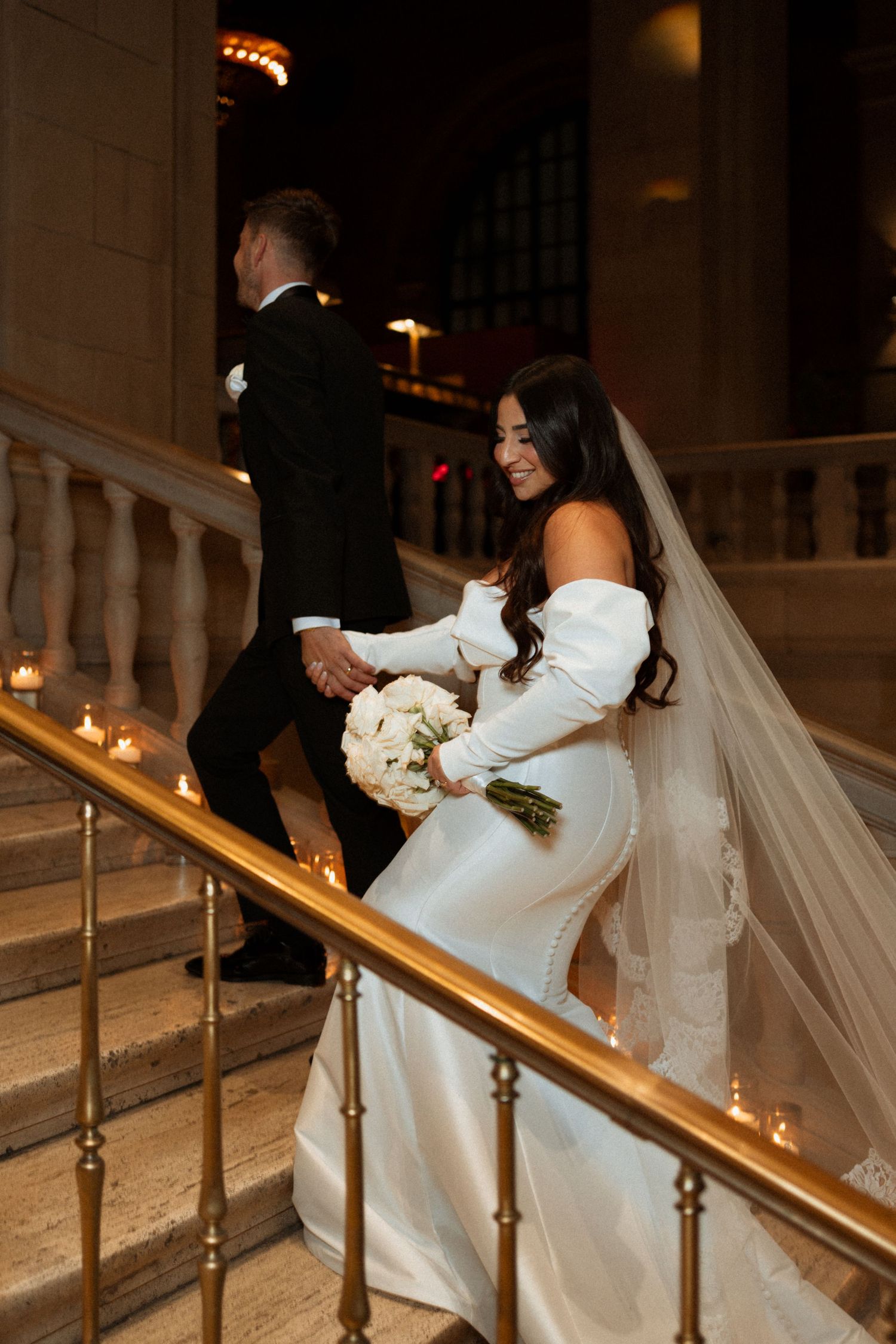 A bride in an elegant off-shoulder gown ascends candlelit stairs during her wedding.