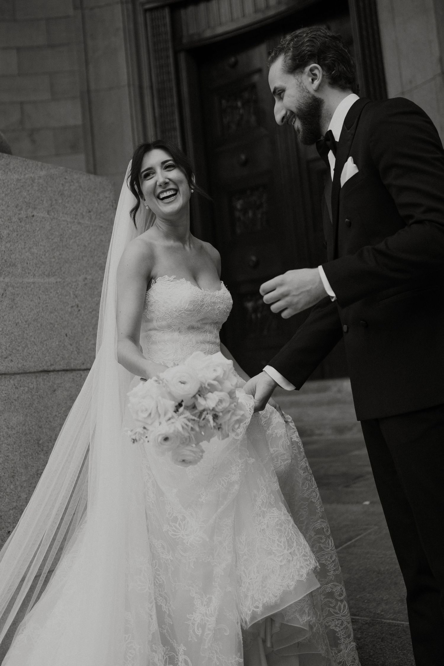 Black and white photo of a laughing bride in lace gown holding bouquet with groom in tuxedo on church steps.