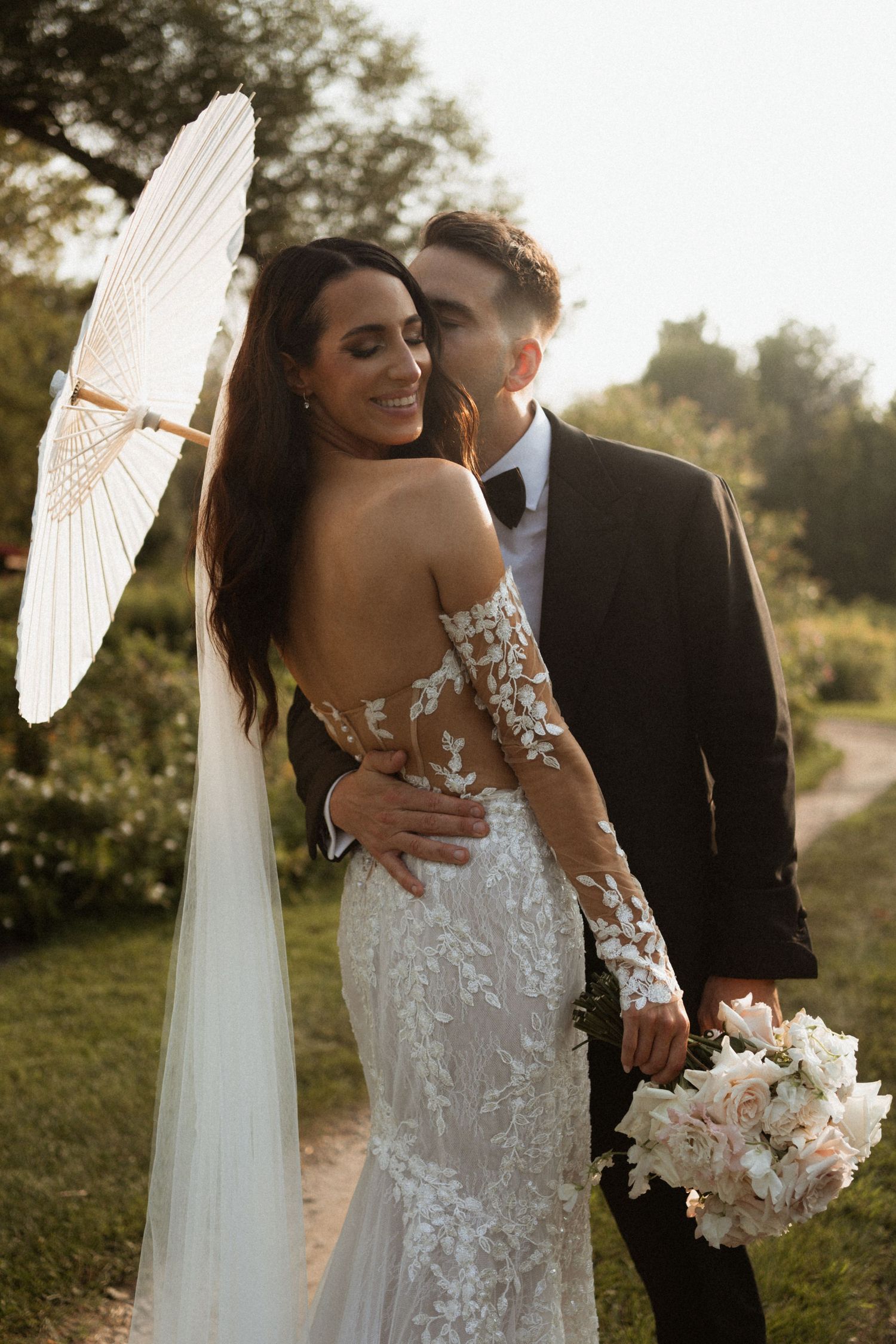 Bride in lace dress holds parasol while groom embraces her in golden sunlight.