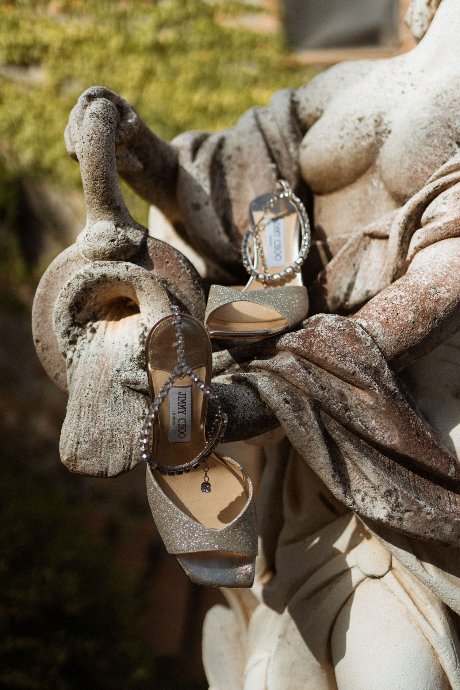 Jimmy Choo glittery heeled sandals displayed on a weathered stone statue in an outdoor garden setting.