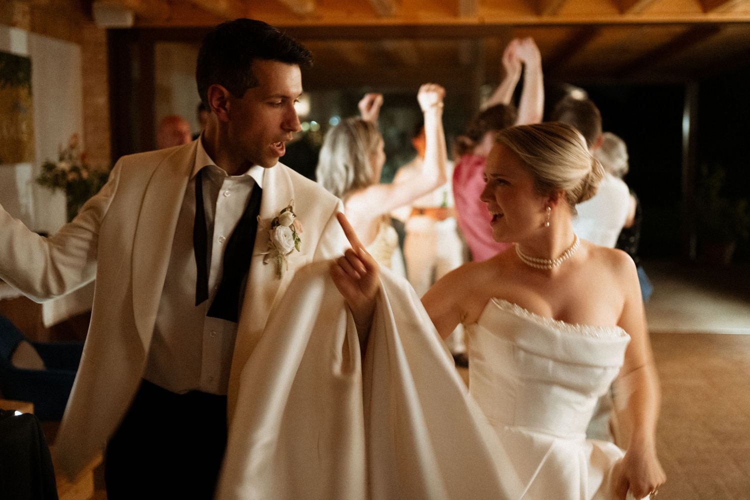 Bride and groom dancing joyfully at wedding reception, bride lifting white gown, guests celebrating in background.
