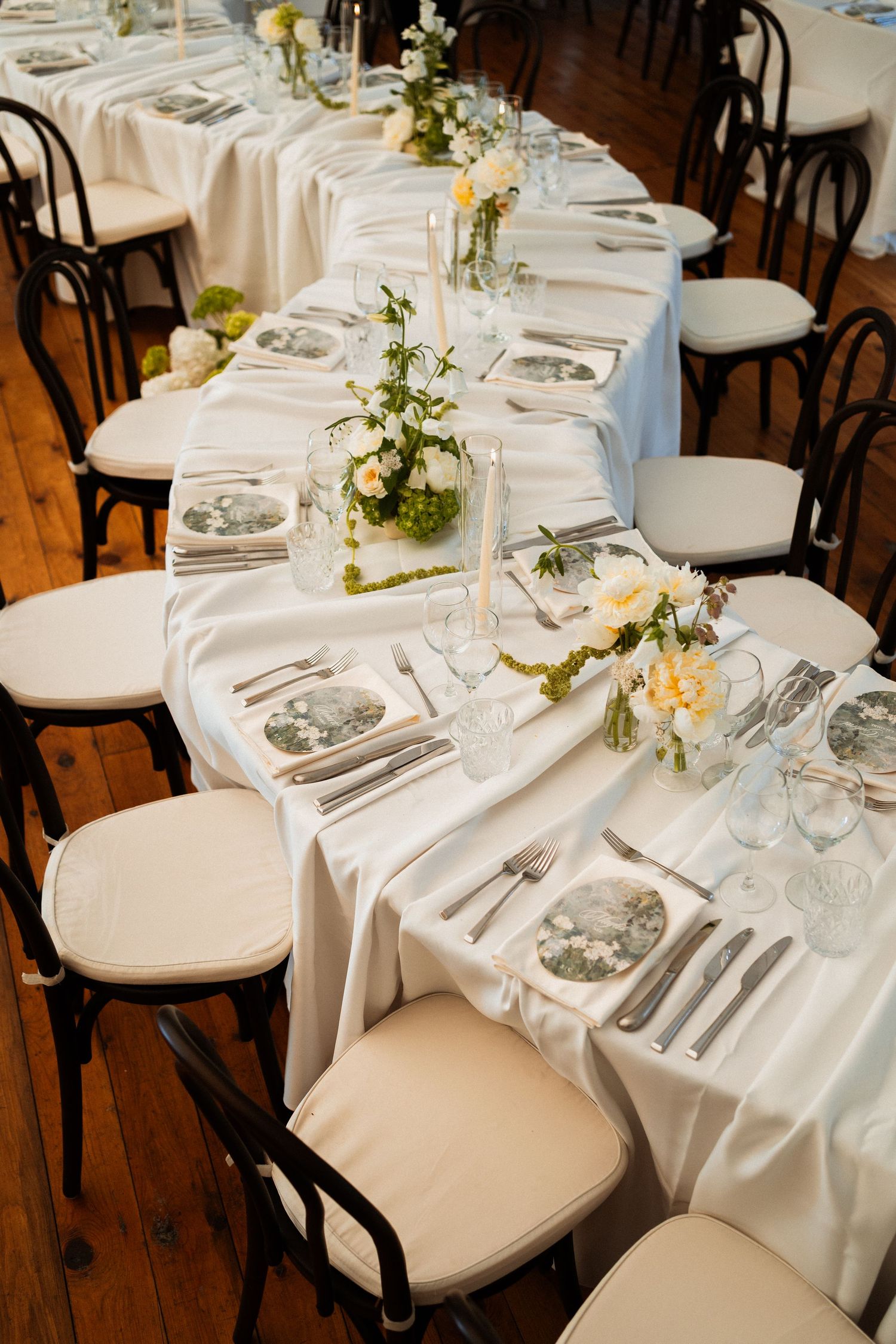 Elegant wedding reception table with white satin runner, floral centerpieces, and black bentwood chairs on hardwood floors.