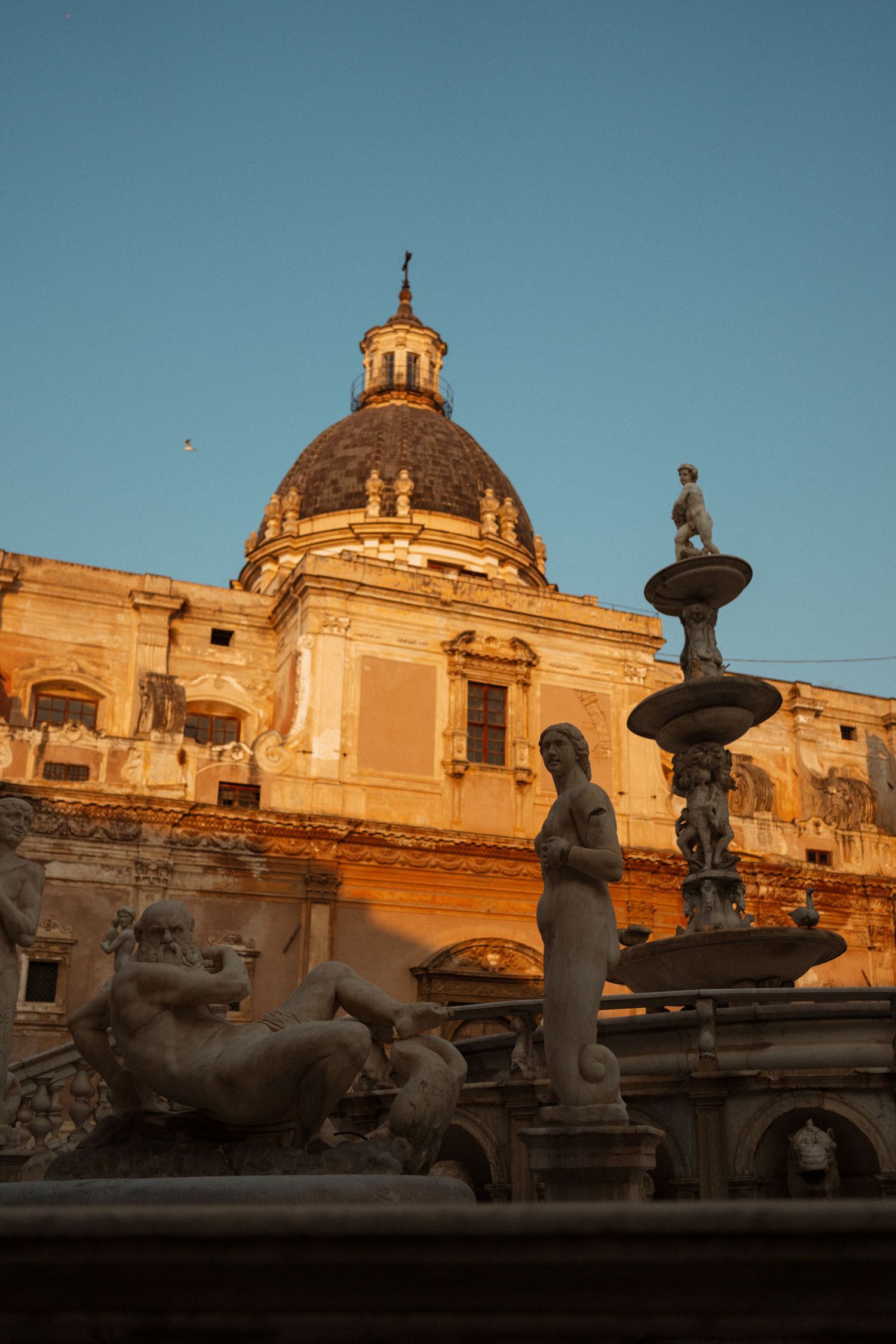 Historic baroque cathedral dome and ornate statues silhouetted against golden sunset light in Rome, Italy.