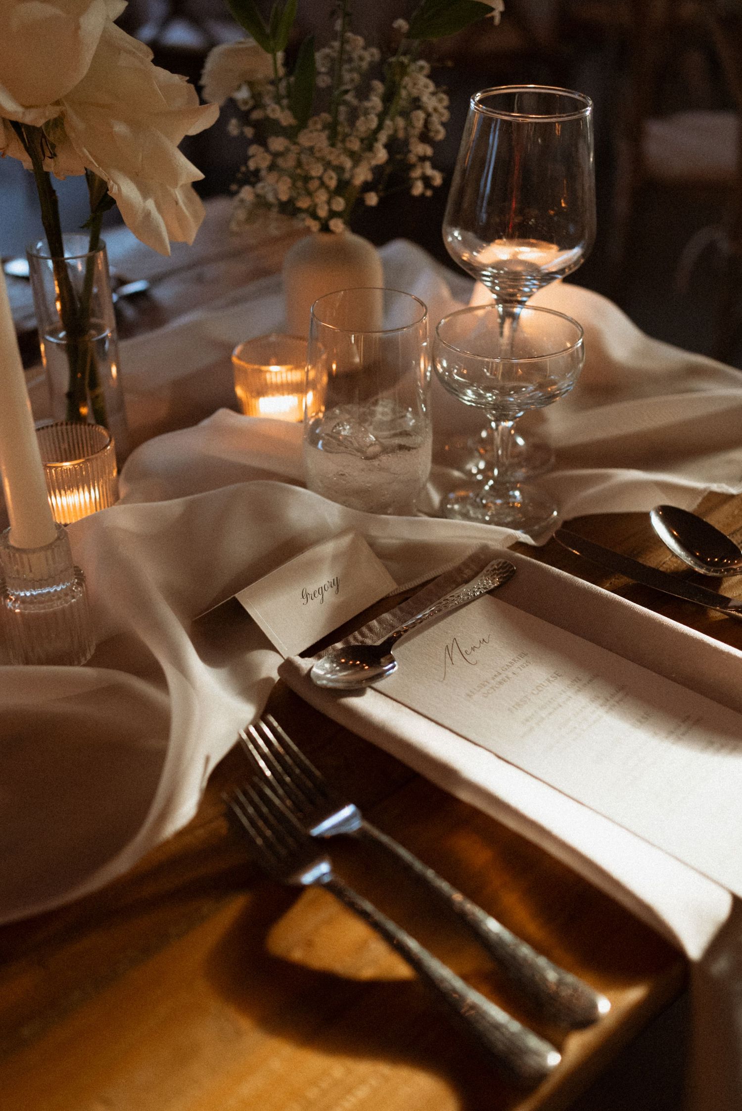 Elegant table setting with wine glasses, candles, and menu cards on white linens in warm ambient lighting.