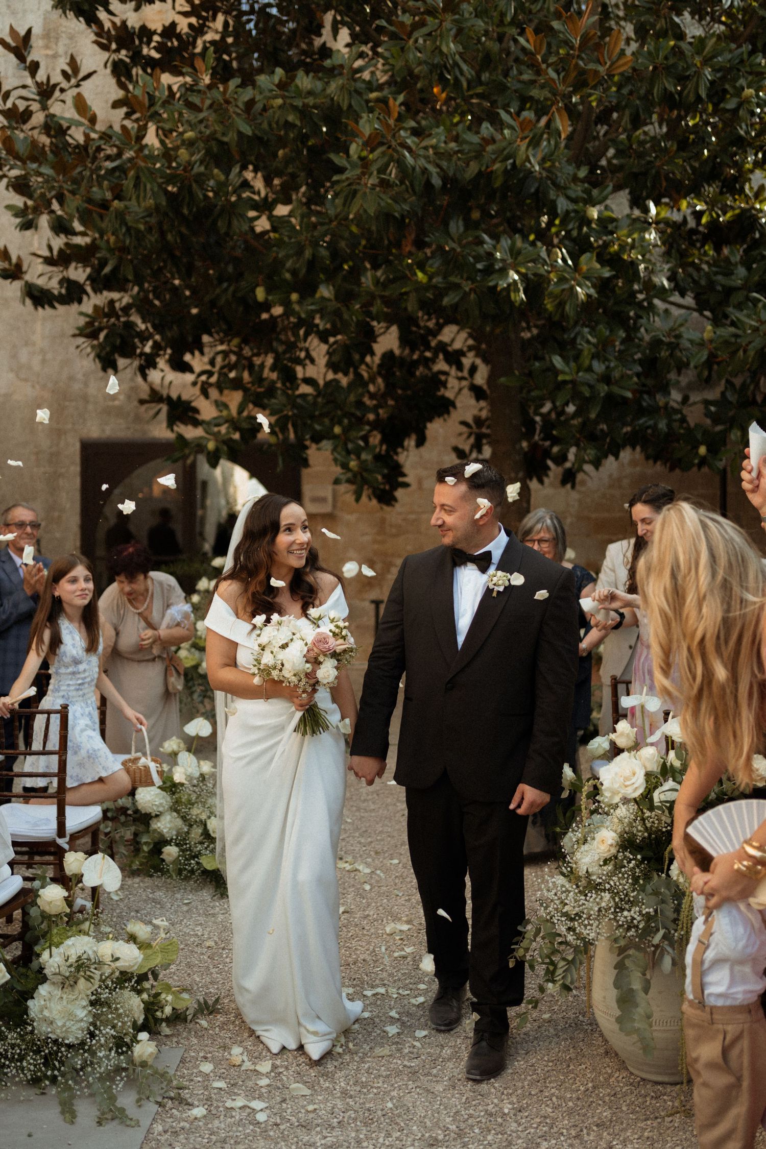 Wedding guests mingling outdoors under trees decorated with white floral arrangements.