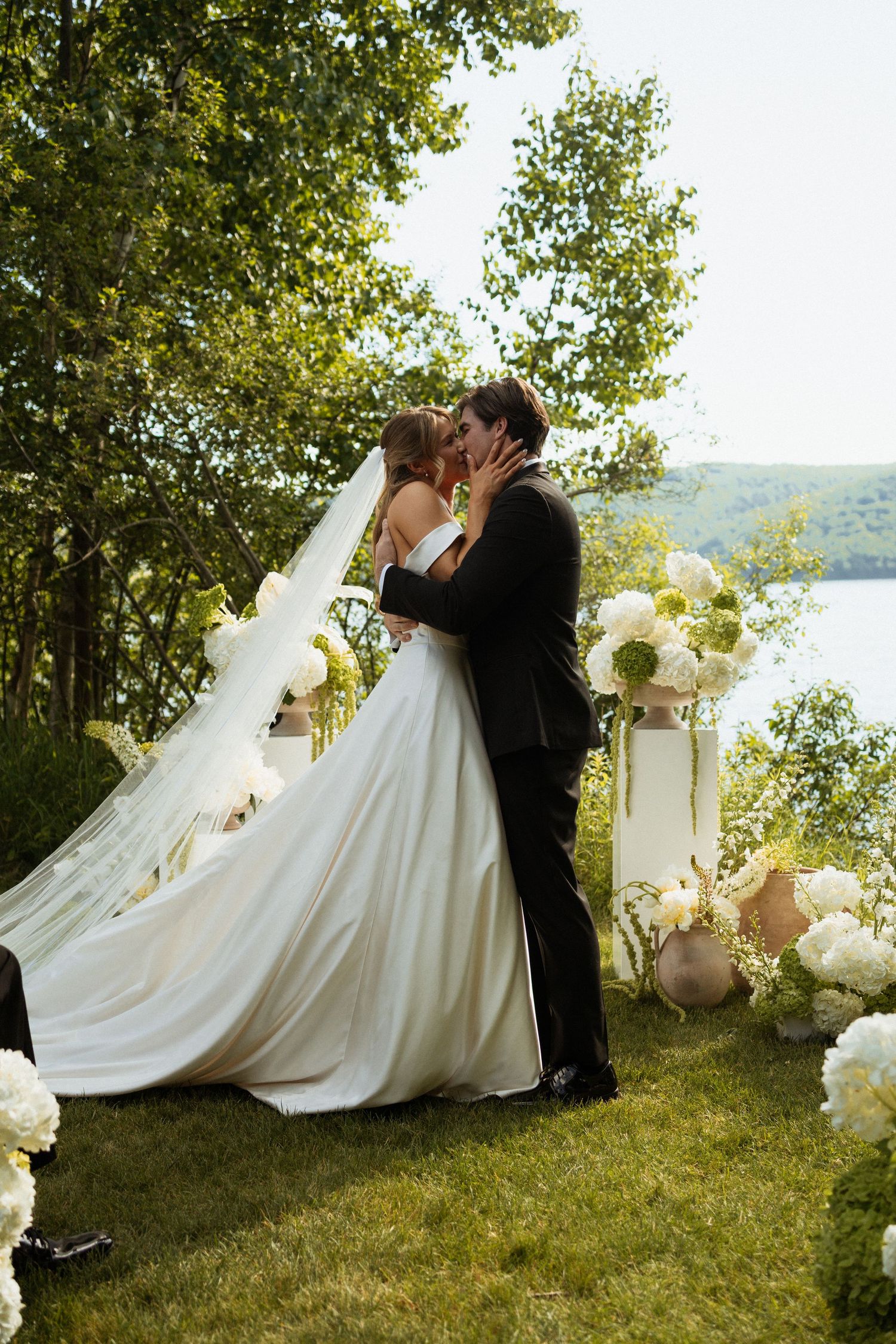 Bride and groom share first kiss at outdoor lakeside wedding ceremony surrounded by white floral arrangements and lush greenery.
