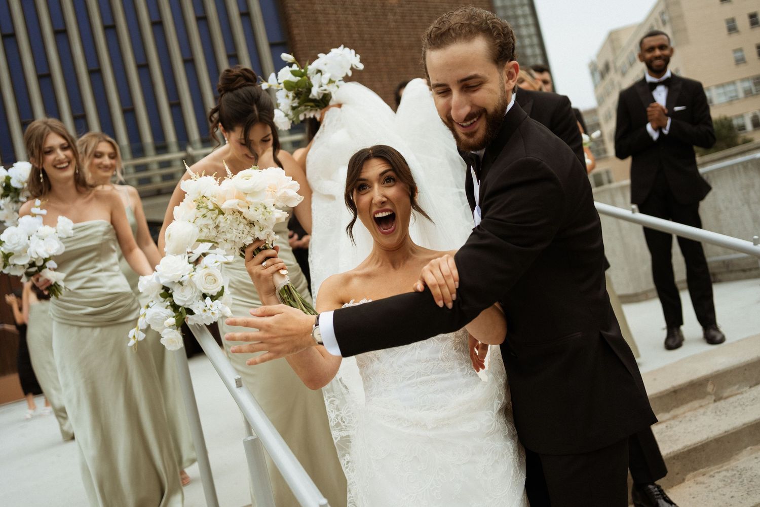 Joyful bride and groom walk together after wedding ceremony, bridesmaids in sage green dresses holding white bouquets behind them.