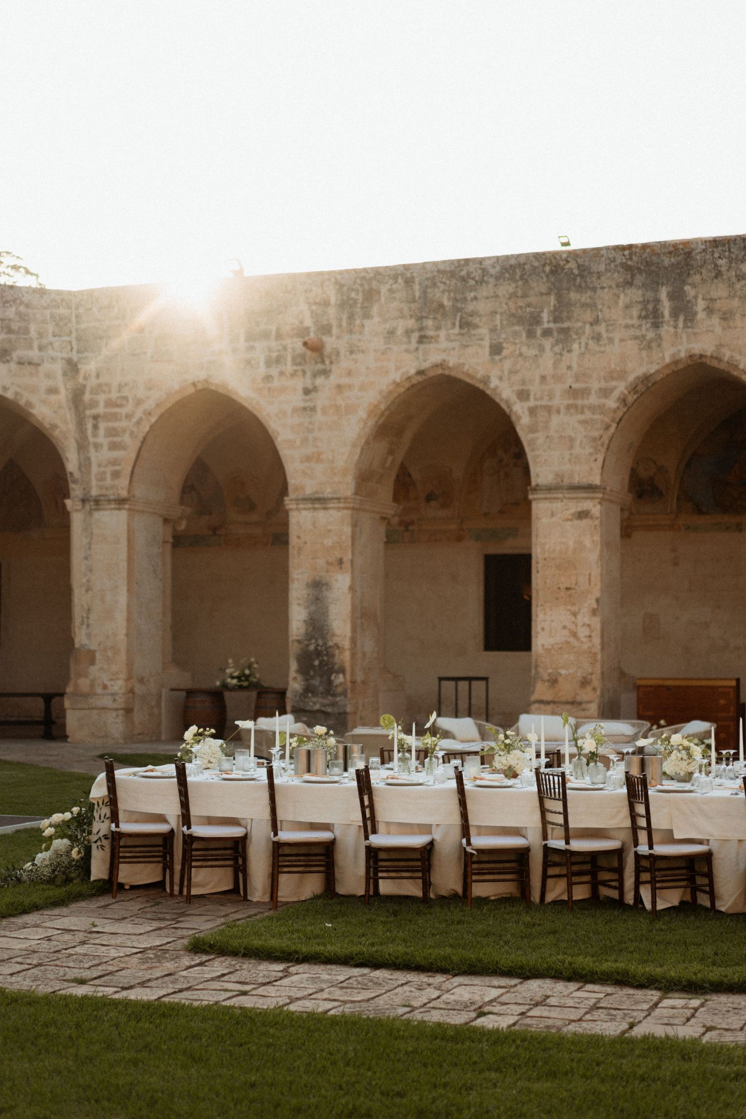 Long rustic dining tables set beneath stone arches in an ancient courtyard during golden hour sunset.