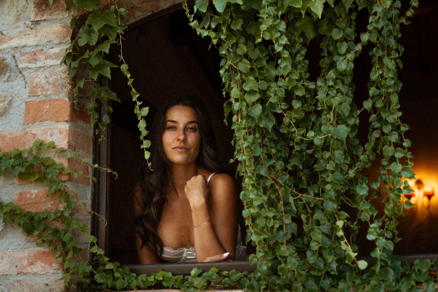 Person in black top poses near ivy-covered brick wall with warm lighting and rustic ambiance.
