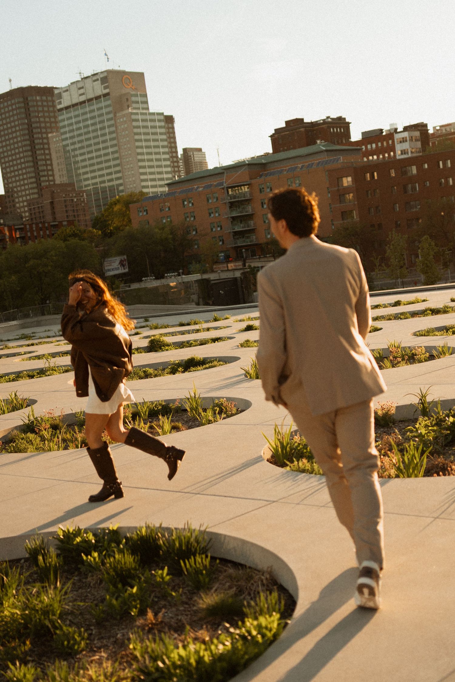 A couple walks through an urban park at golden hour, with city buildings in the background and curved garden paths.