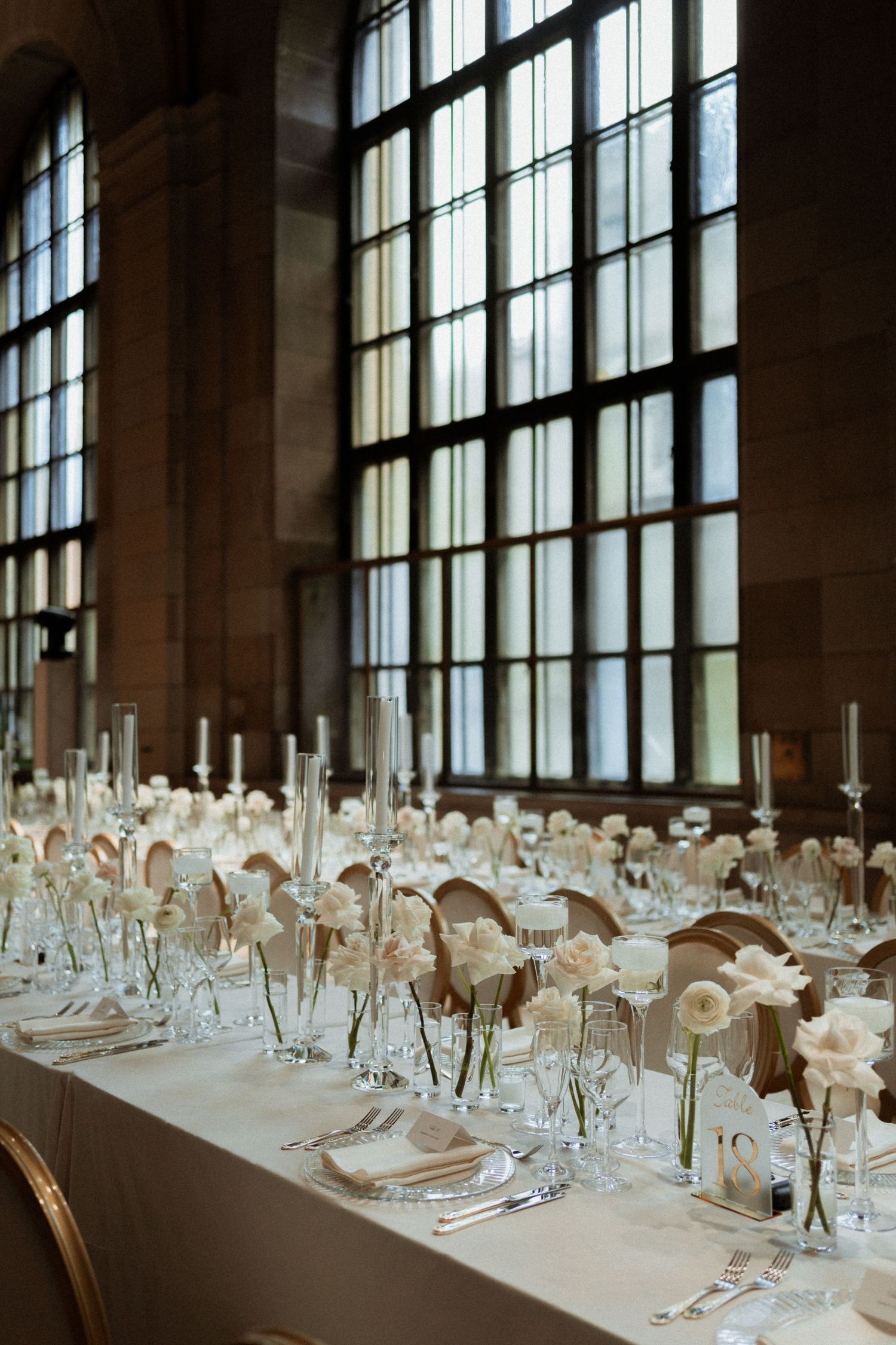 Long banquet table set with elegant glassware and white linens beside tall windows in a bright dining room.