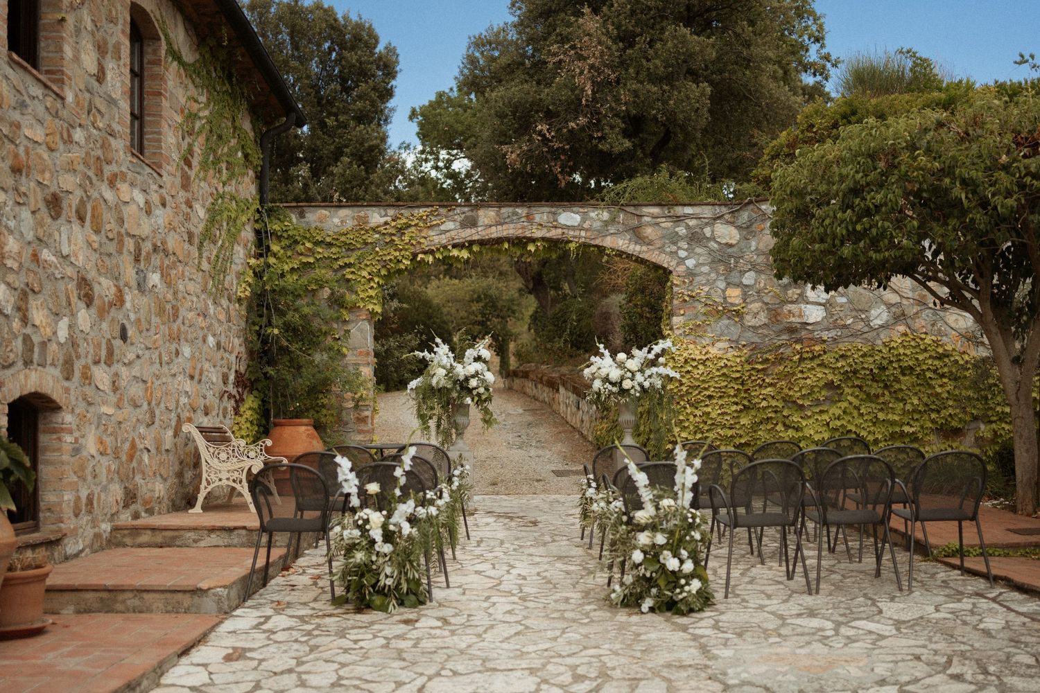 Stone pathway lined with white floral arrangements leads through rustic courtyard at outdoor wedding venue.