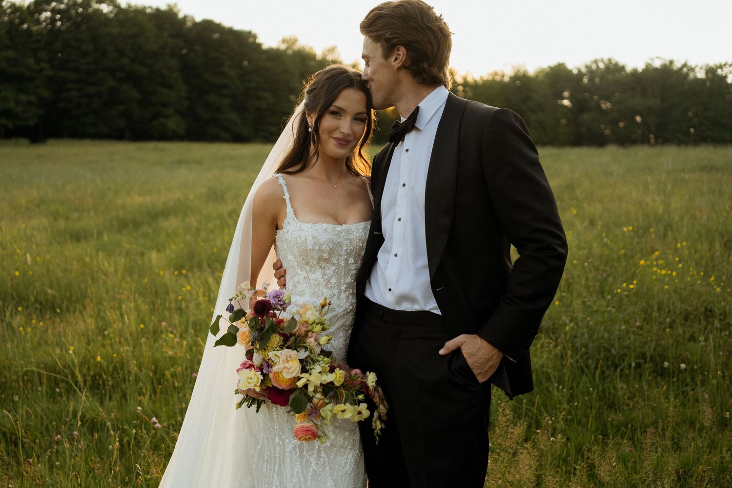 Bride and groom standing in a sunlit green field, bride holding a colorful bouquet, groom in black tuxedo kissing her cheek.