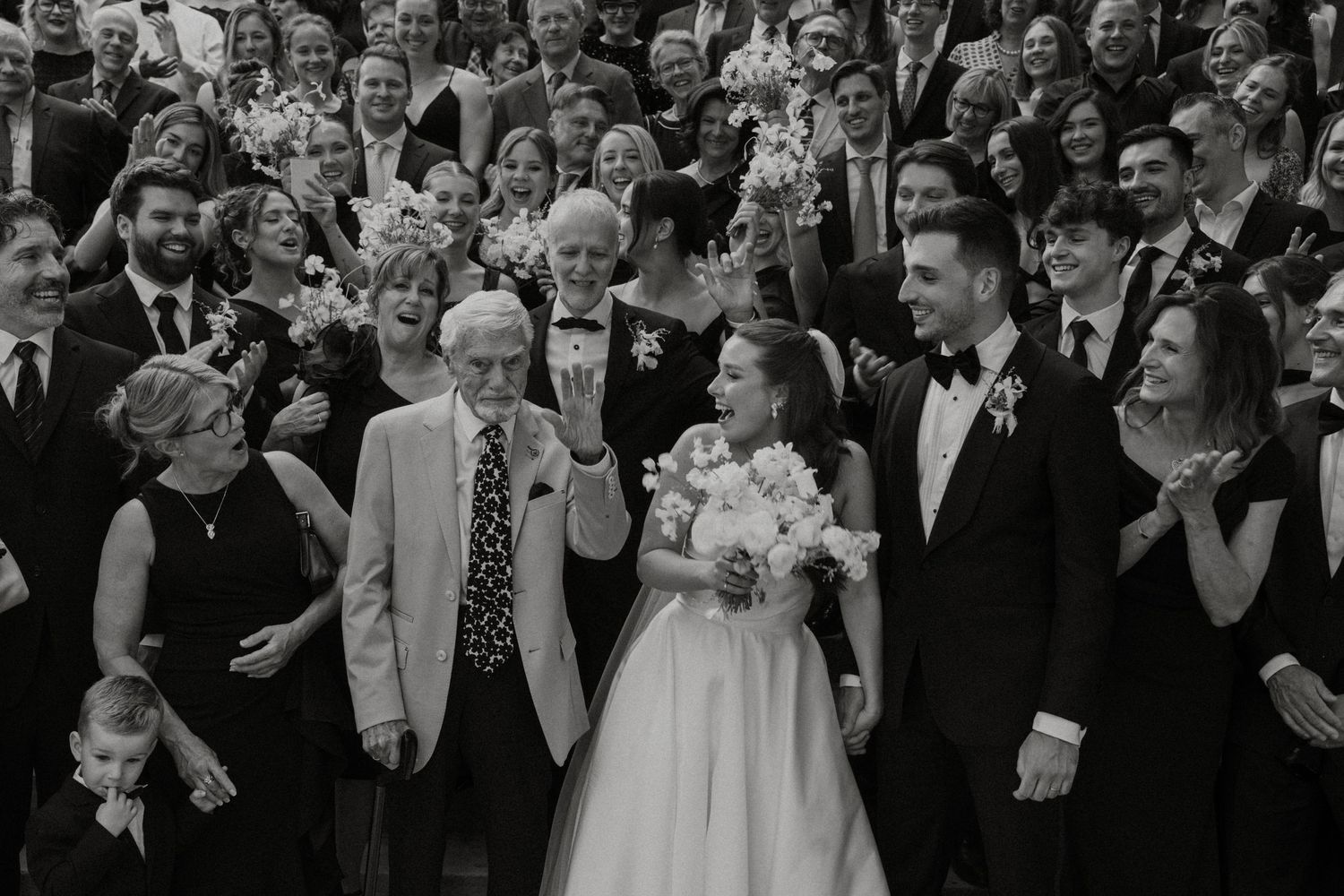 Wedding guests gather outdoors in formal attire while celebrating and taking photos at a joyous celebration.