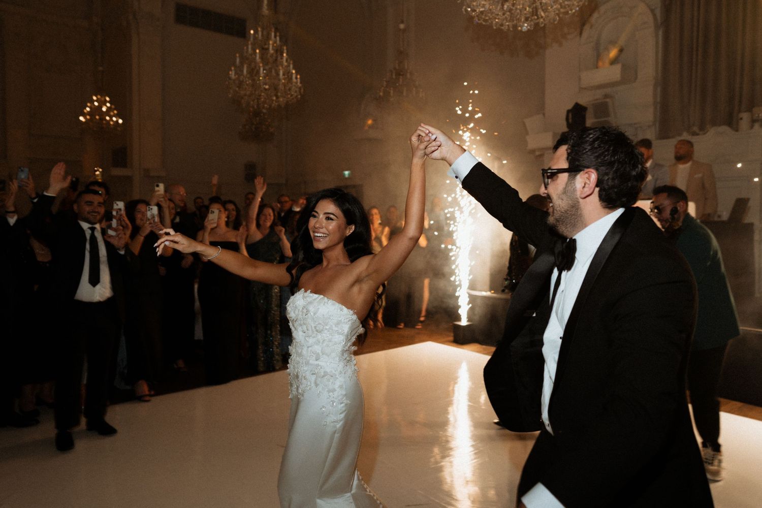 A couple shares their first dance at an elegant wedding reception under a crystal chandelier while guests look on.