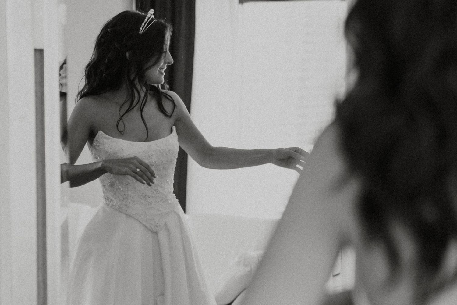 A bride in a white wedding gown stands by a window in a black and white photograph.