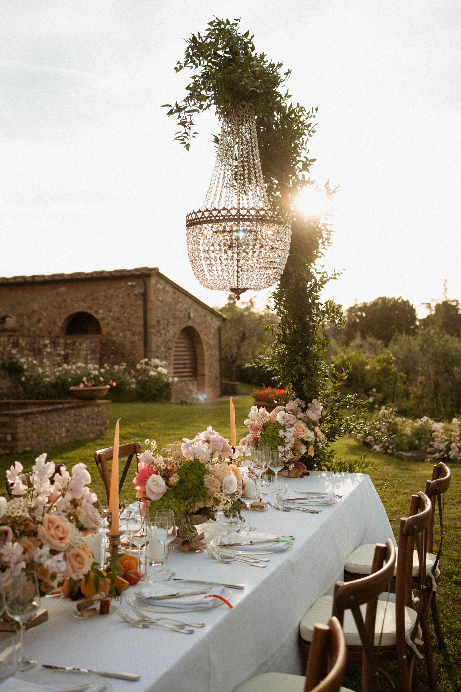 Elegant outdoor wedding reception table with crystal chandelier, blush floral arrangements, taper candles, and Tuscan villa backdrop.