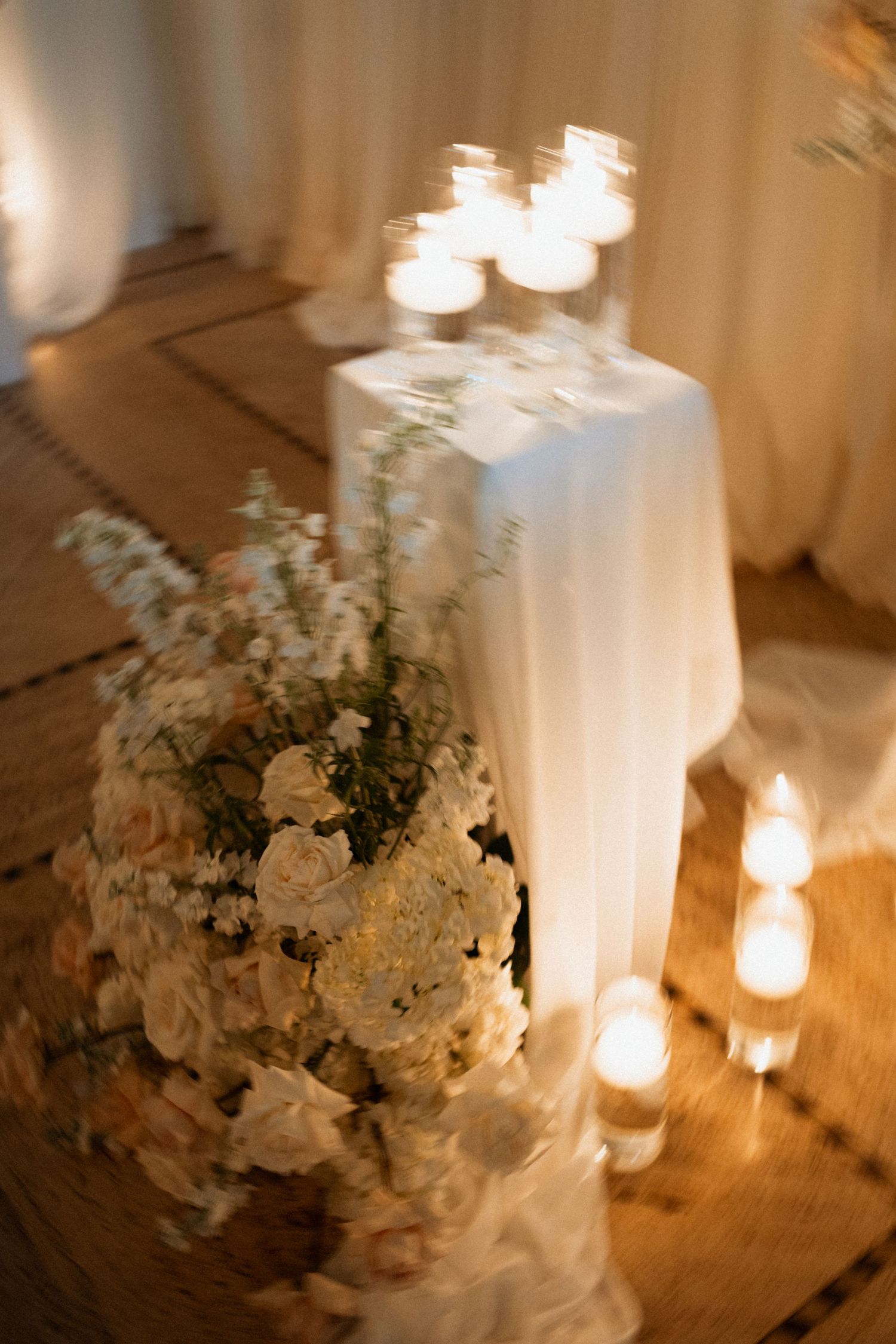 A white floral wedding centerpiece arrangement with glowing candles creates a romantic ambiance on a wooden table.