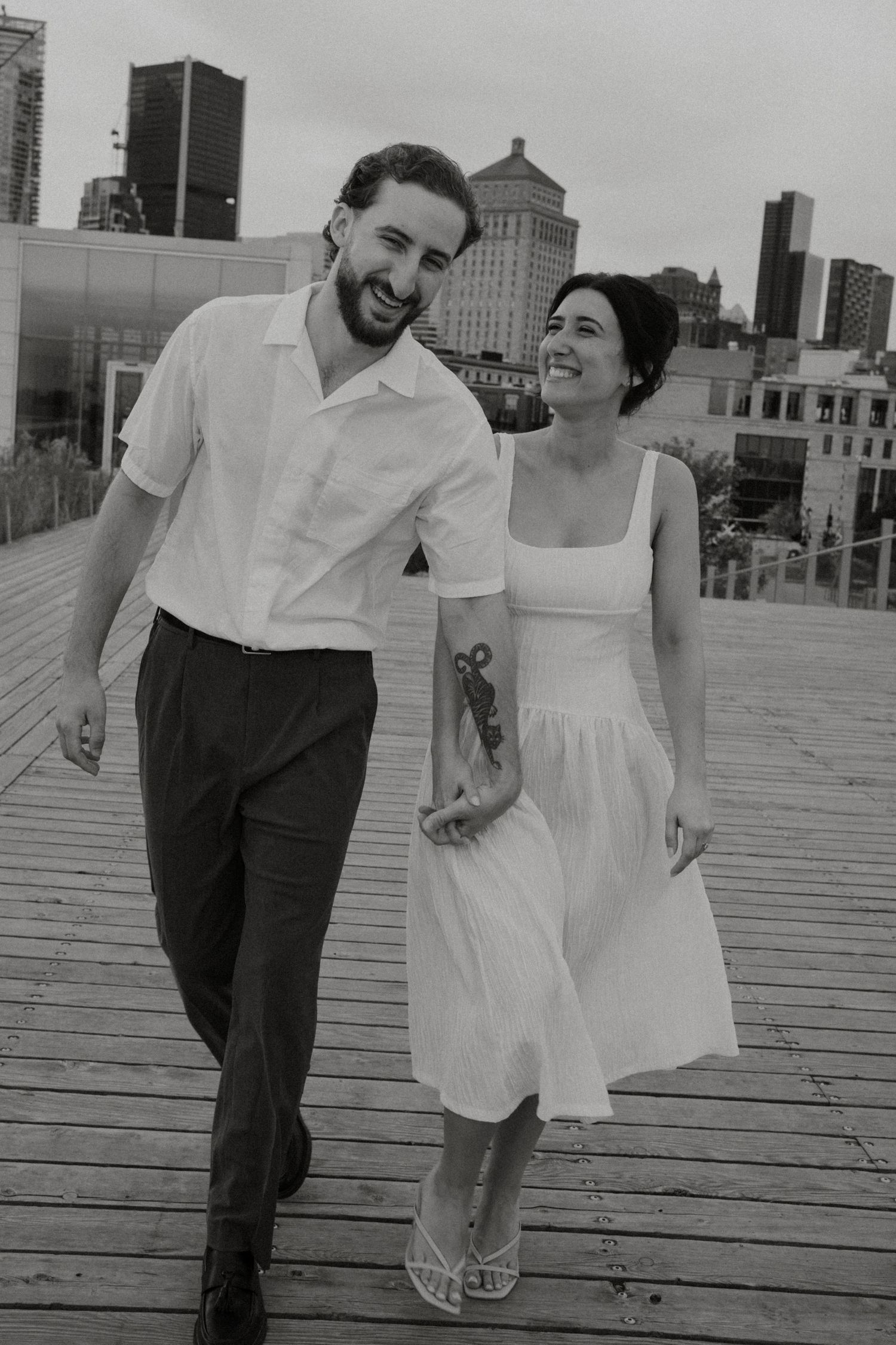 Couple walking together and laughing on urban rooftop with city skyline in background.