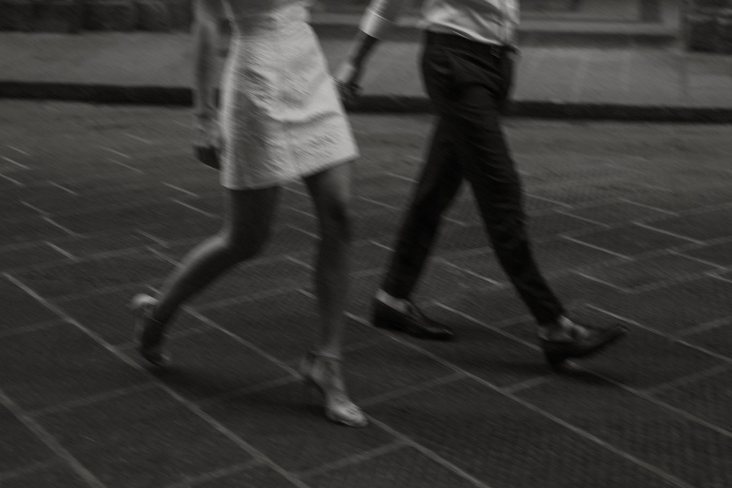 Blurred black and white photo of two people walking on a tiled sidewalk, capturing motion in urban setting.
