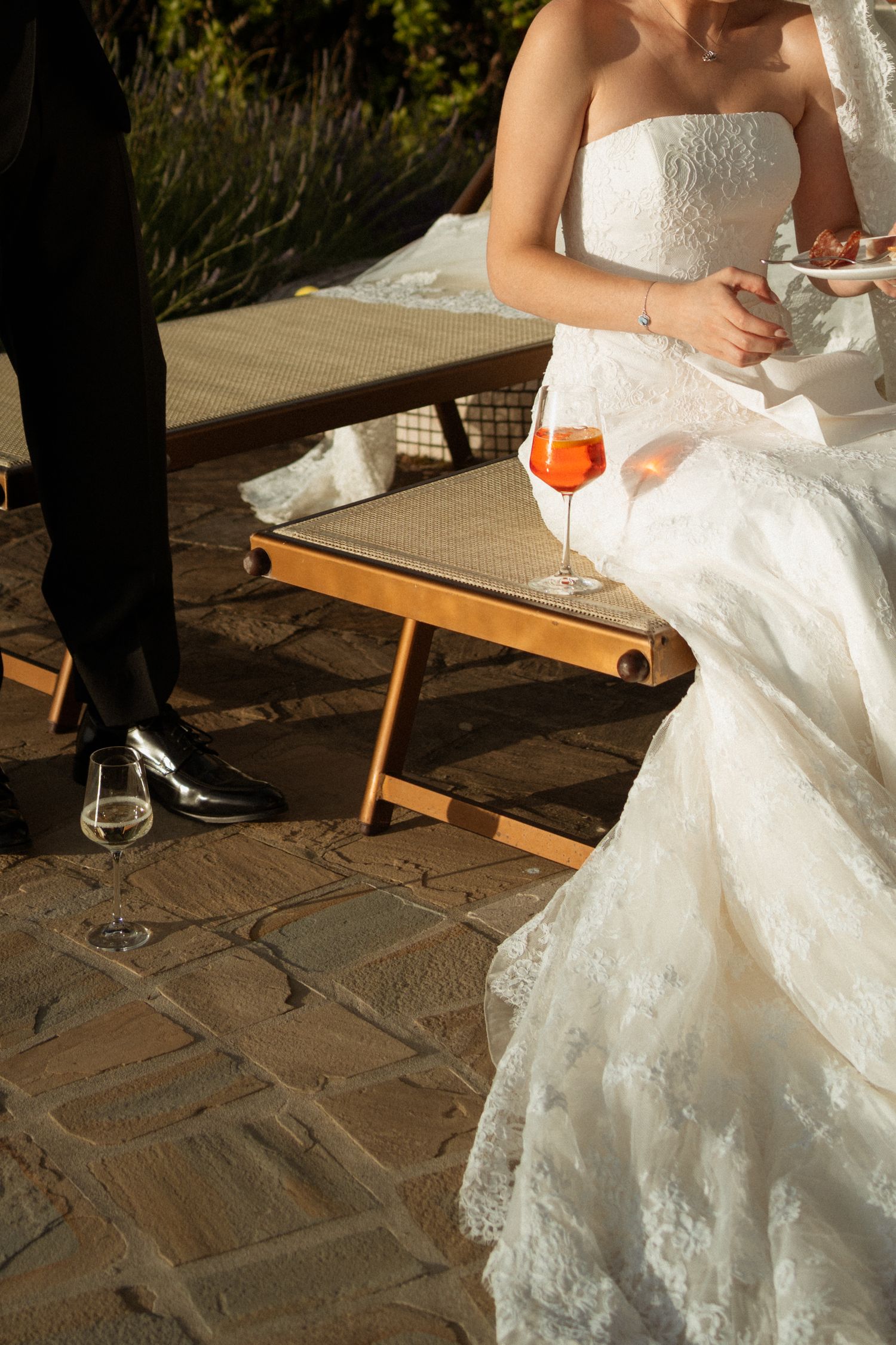 Bride in white lace gown sitting outdoors with an Aperol Spritz cocktail during a wedding reception.