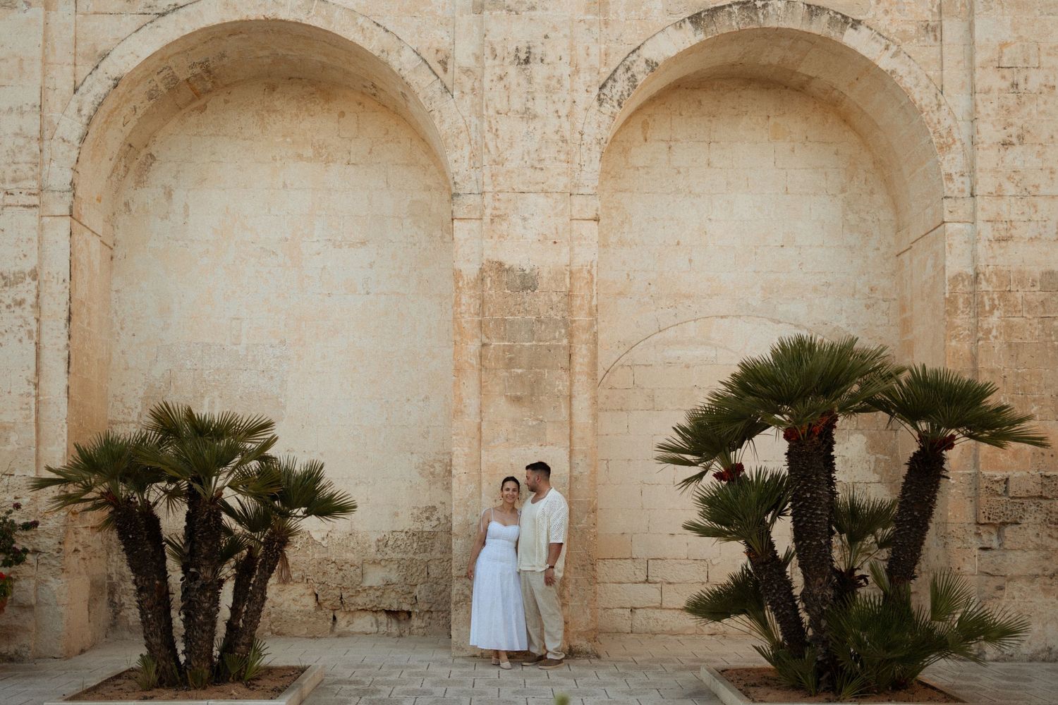Couple standing between palm trees against stone archways of historic Mediterranean building at sunset.