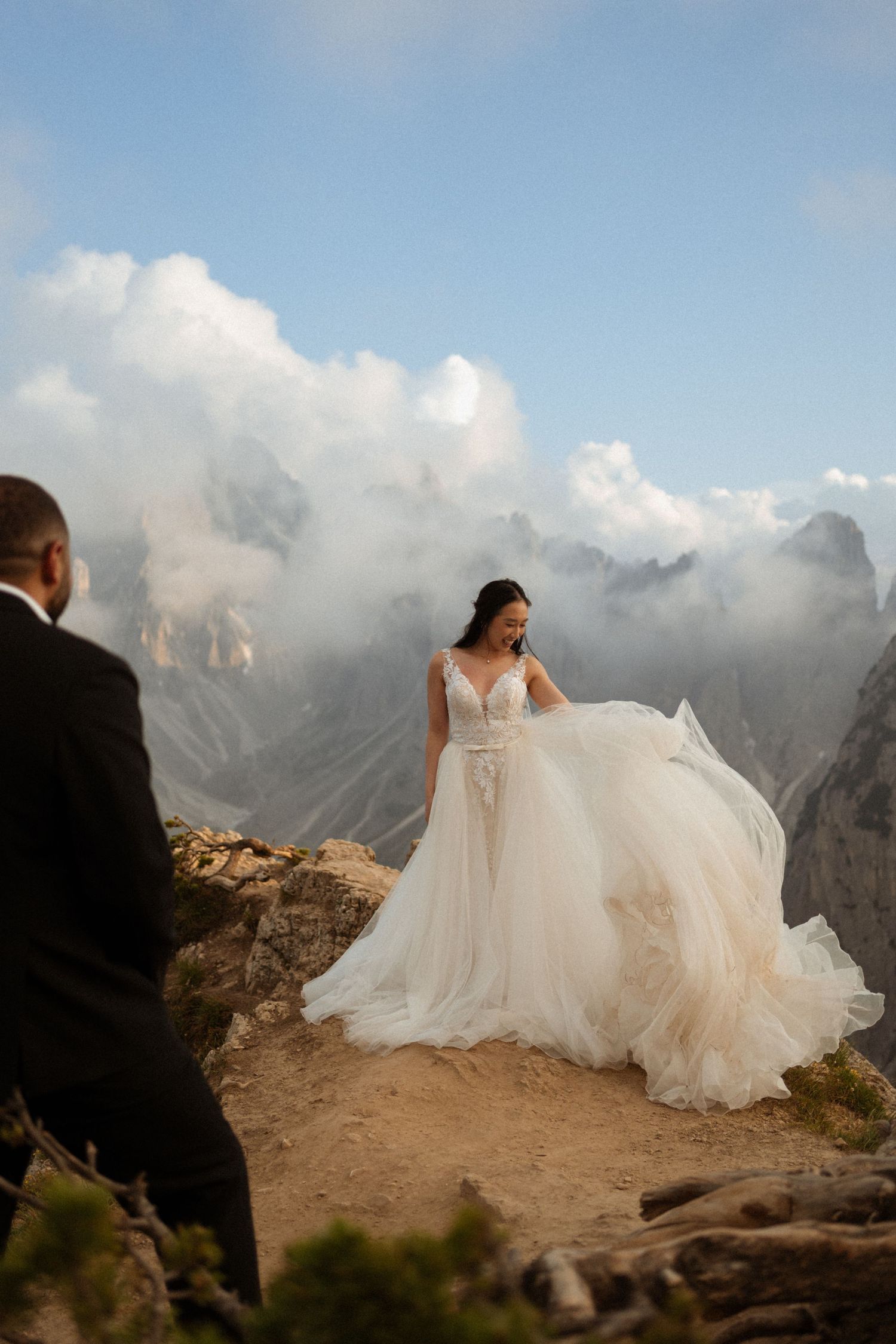 Flowing white wedding dress billows dramatically against a mountainous landscape with clouds.