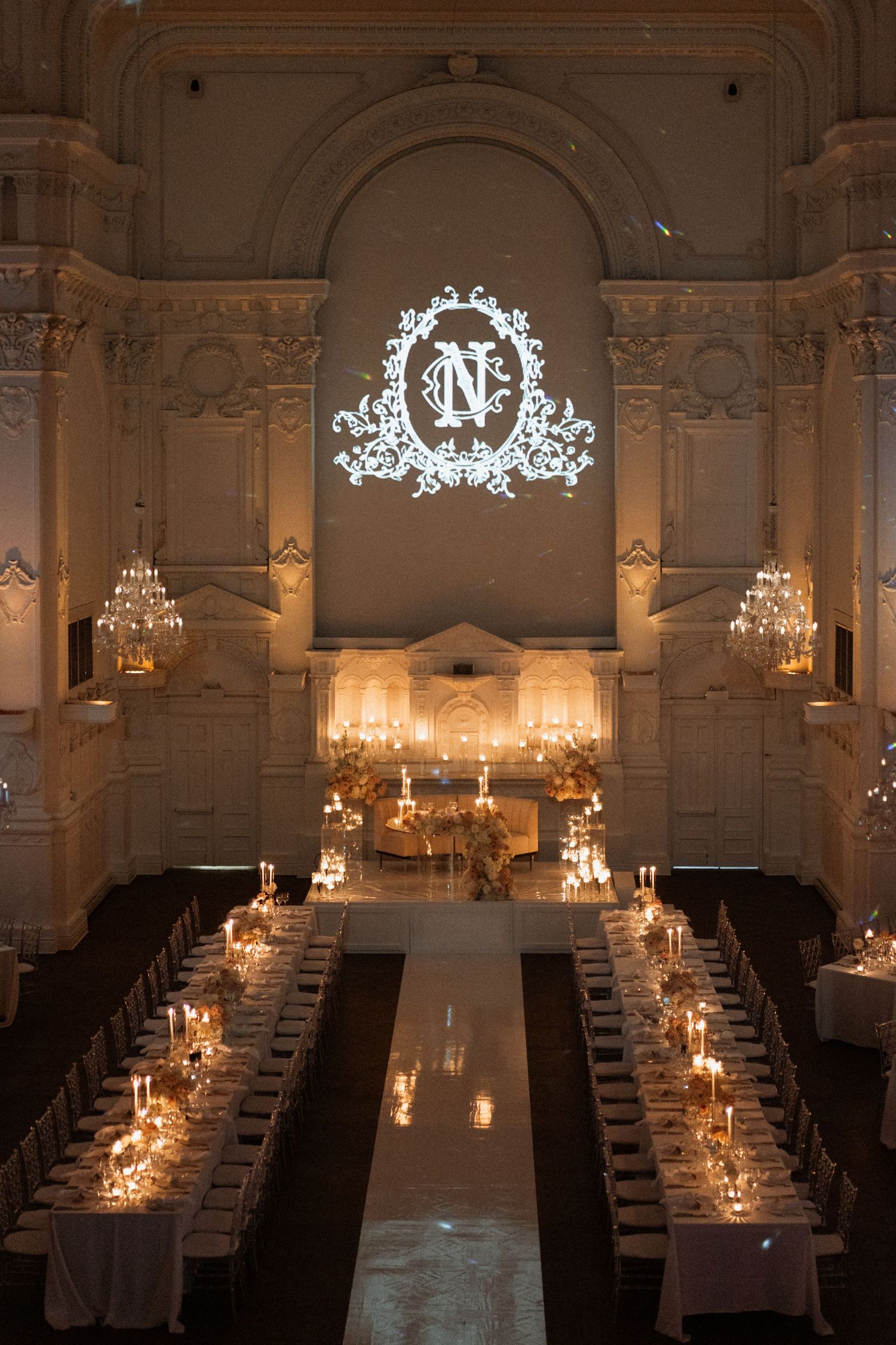 Elegant wedding reception hall with long tables, crystal chandeliers, and illuminated monogram projection on arched wall.